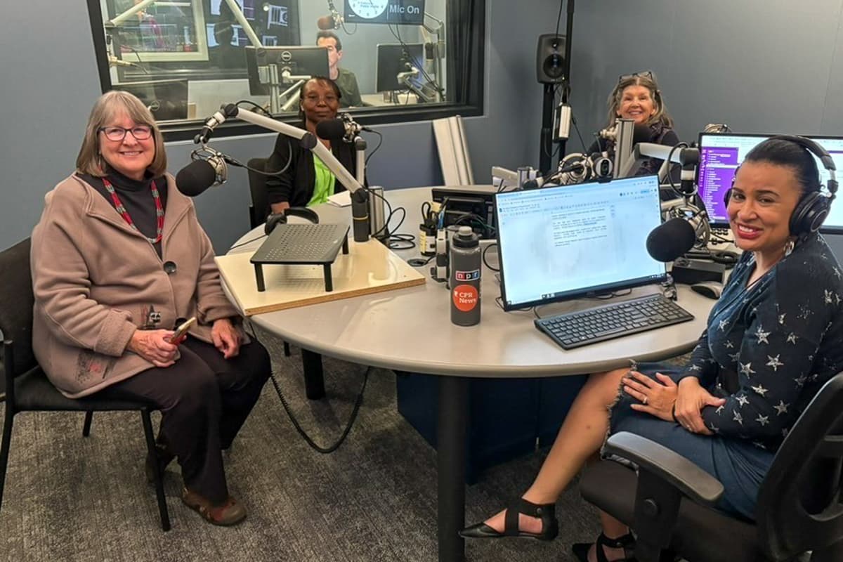 Image shows four women in a studio seated at microphones around a round table.