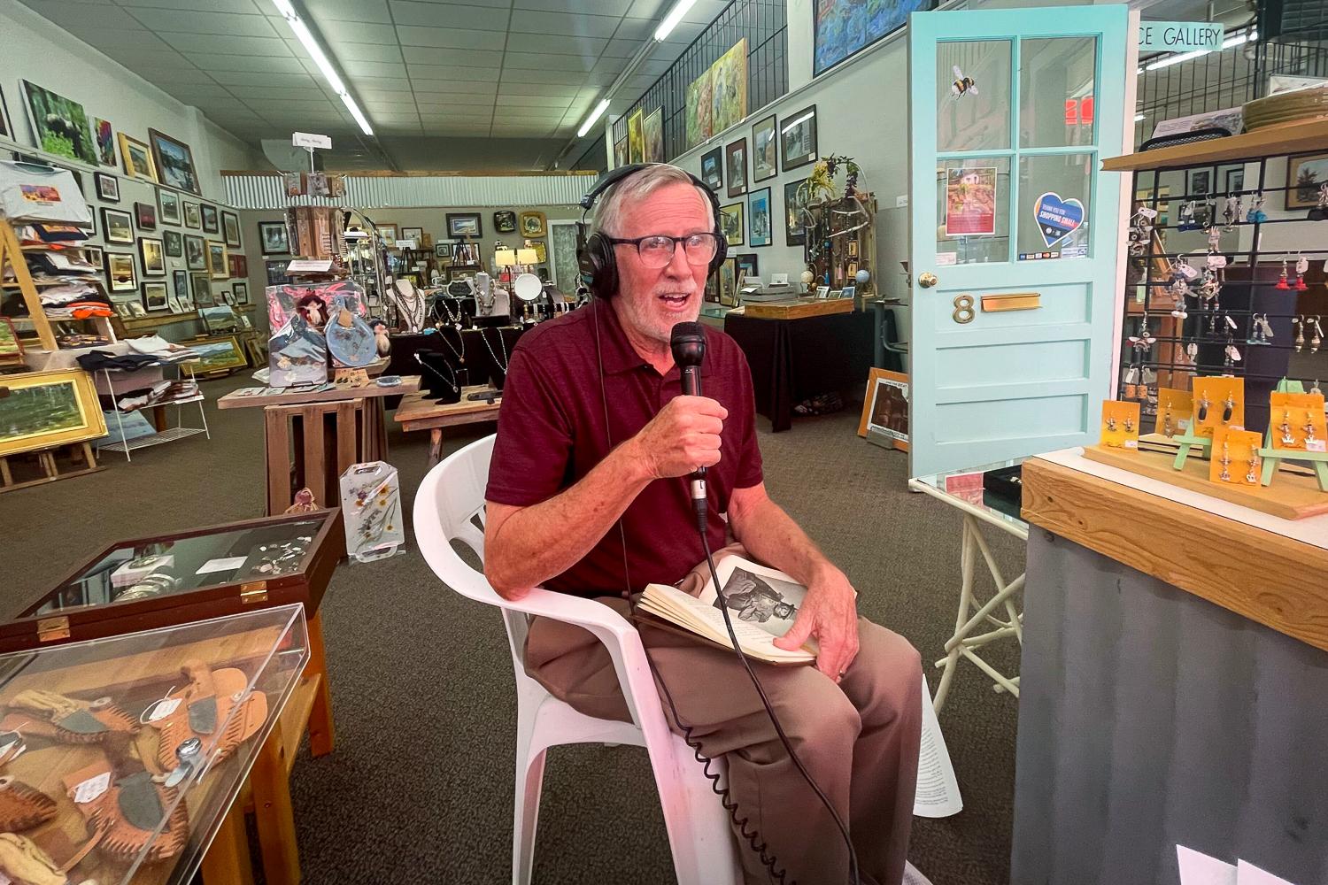A man sits in a chair holding a microphone with headphones on and a book in his lap. He is in a arts and crafts store.
