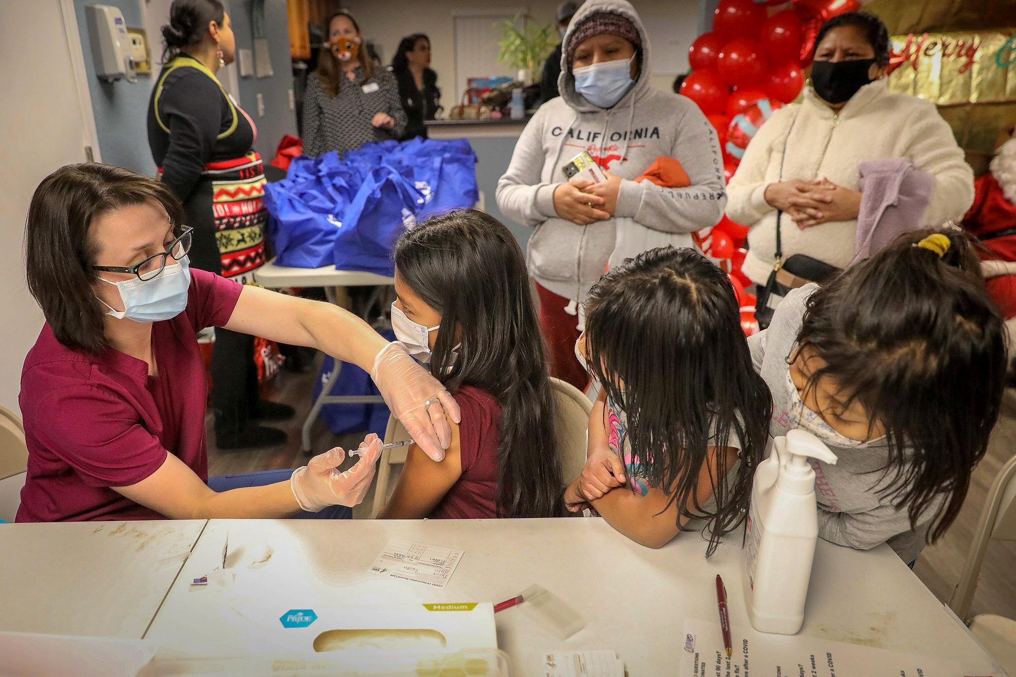 Three children sit at a table while one of them receives a shot