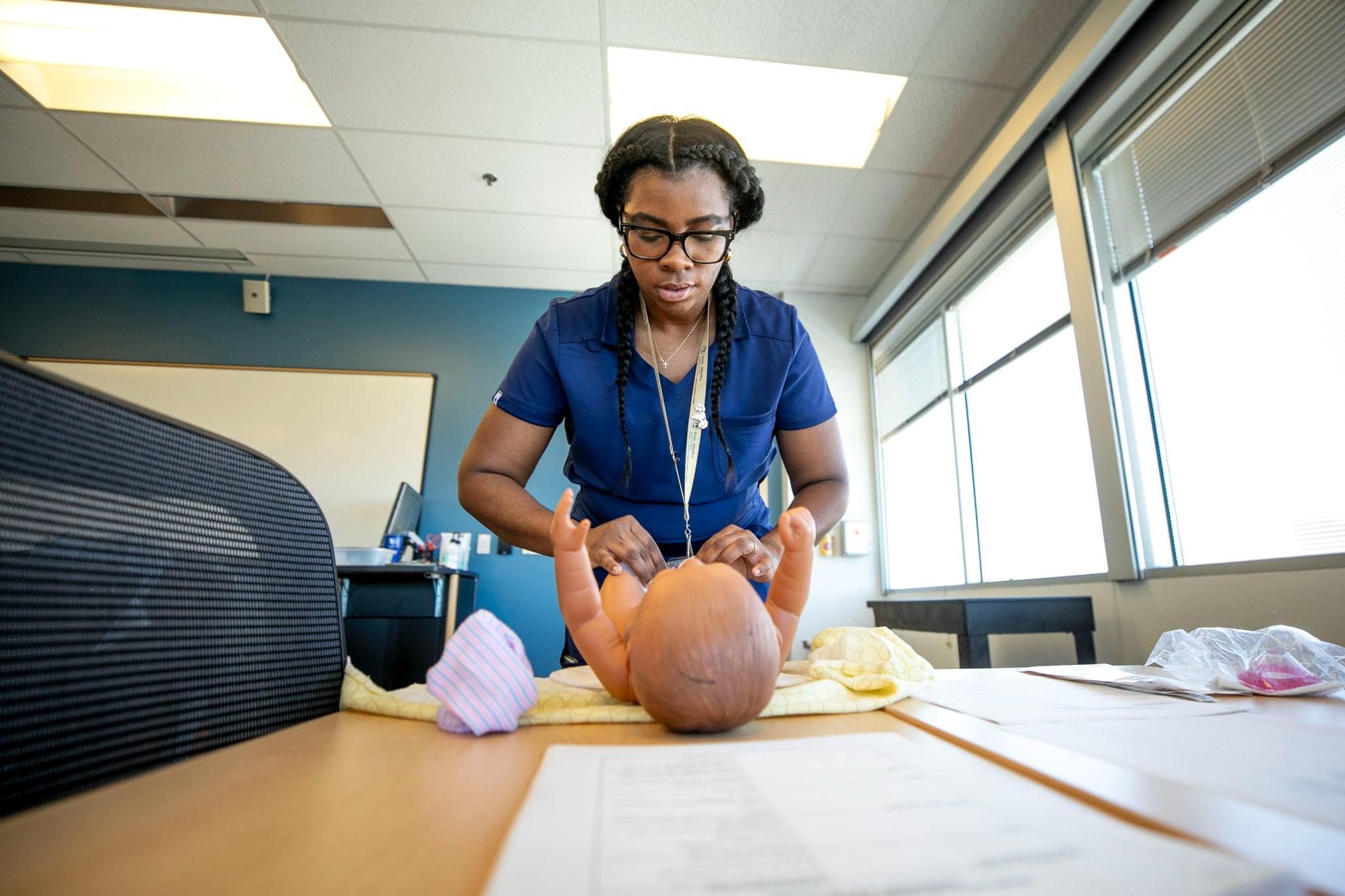 A nurse wearing blue scrubs stands over a baby mannequin