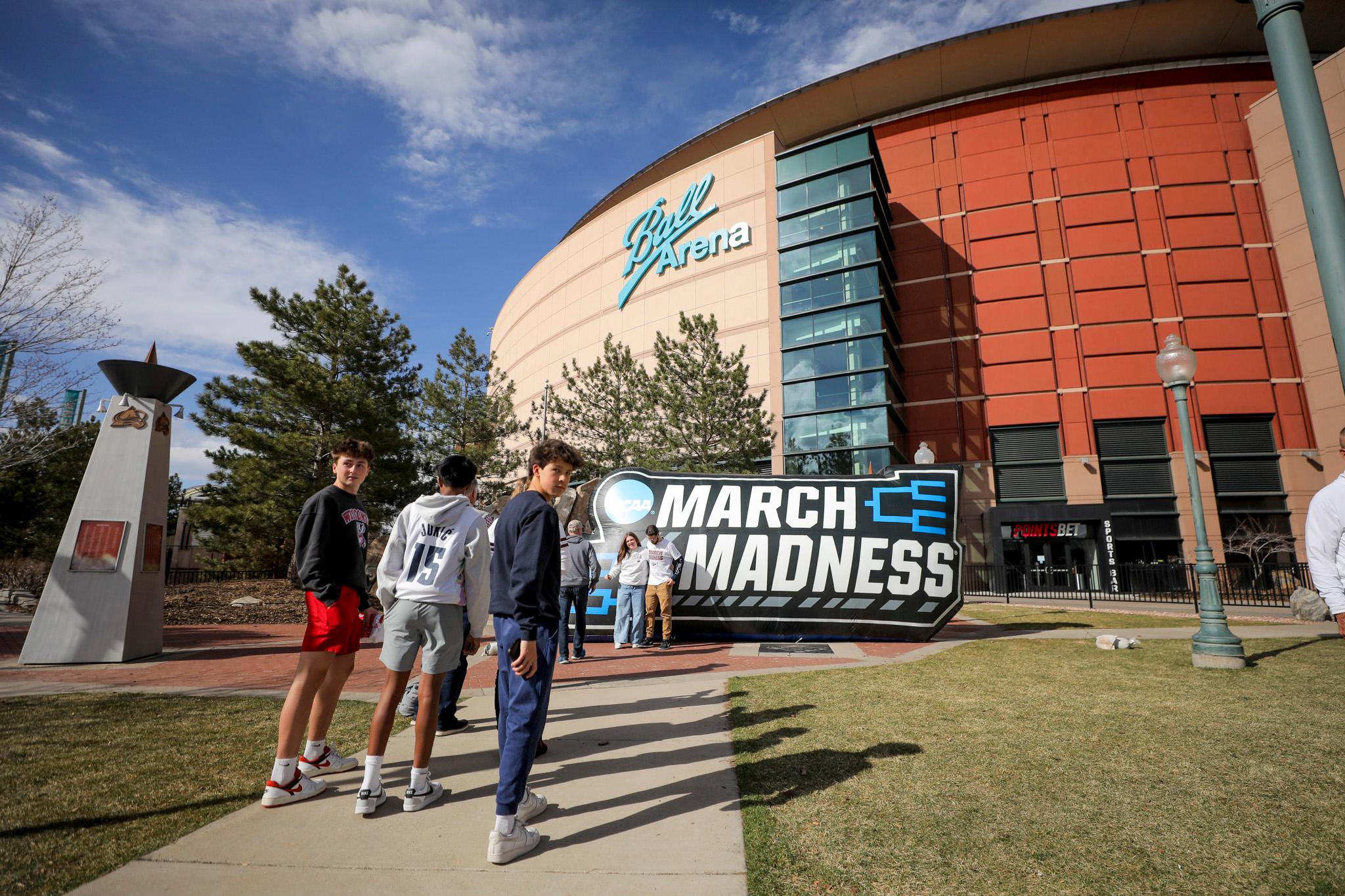 A group of people are standing outside of a Ball Arena in Denver with a large sign that says "March Madness." They are in front of the sign, which is located on the sidewalk.