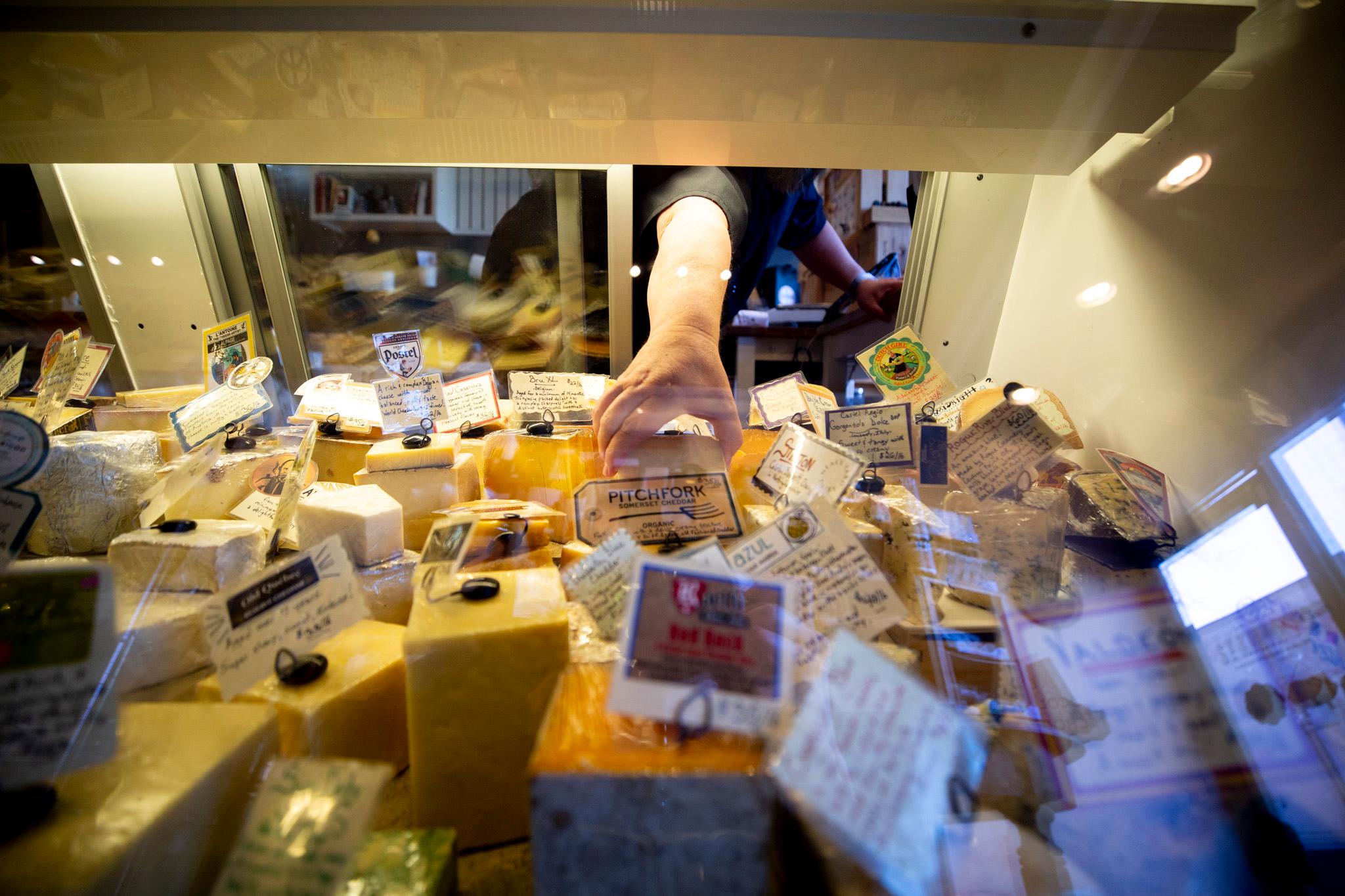 A person is reaching for a piece of cheese in a display case. The cheese is placed on a shelf surrounded by other cheeses.