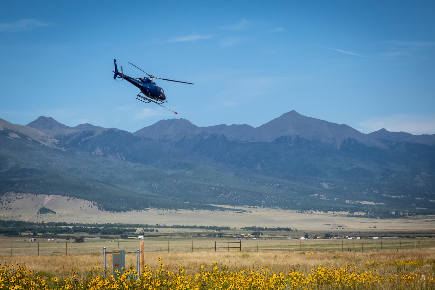 A helicopter flying over a large, open area with mountains in the background and sunflowers in the foreground.