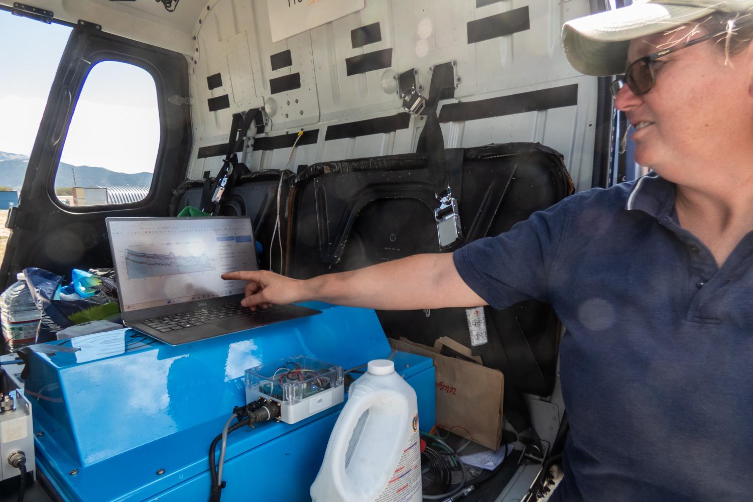 A person is pointing at a laptop screen, which is placed behind the pilots seat in a helicopter. The laptop is open and displaying information. The person appears to be explaining or discussing the content on the screen.