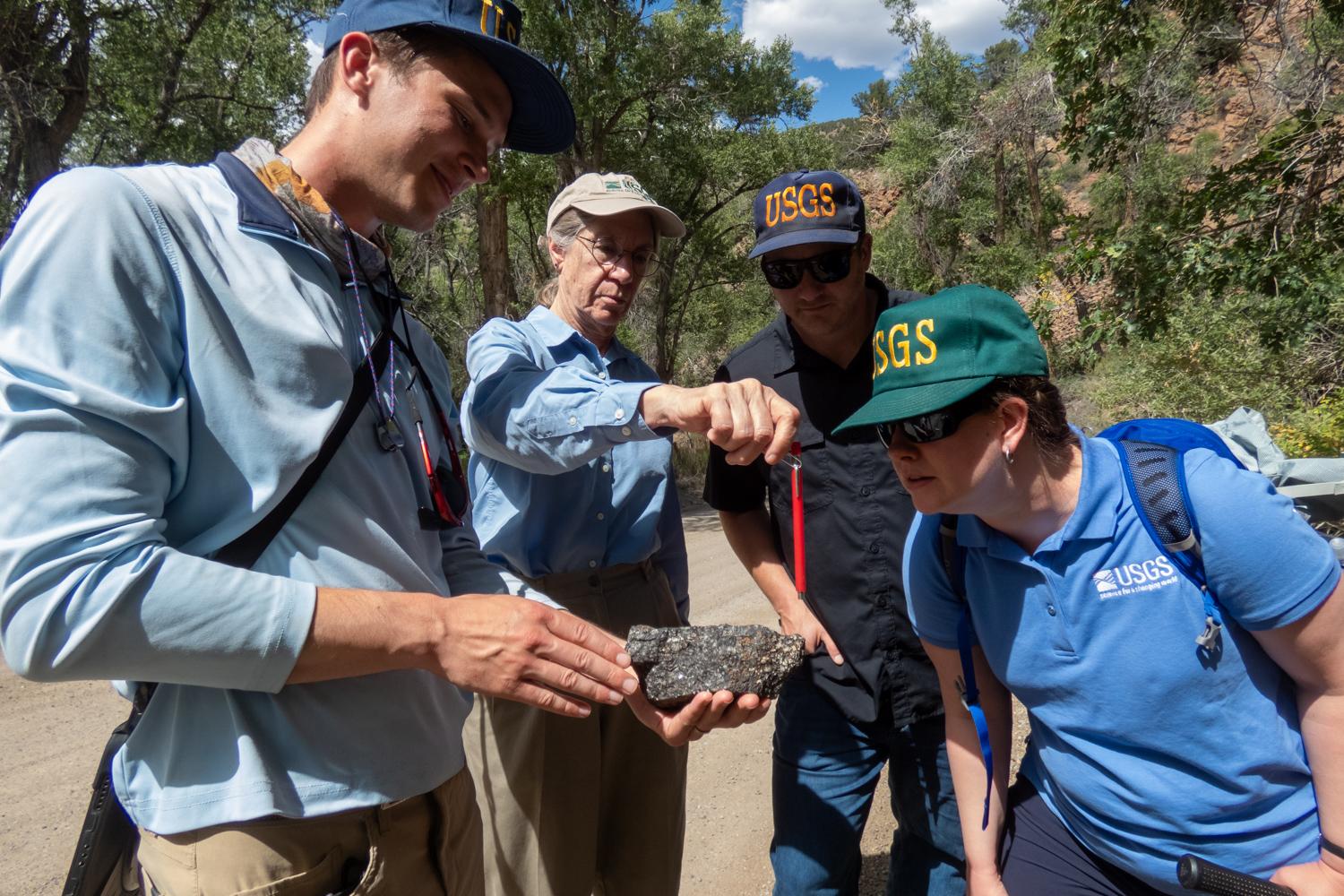Two women and two men examine a black colored rock held in the hand of the man at the left. The scene is outdoors, in a natural setting.