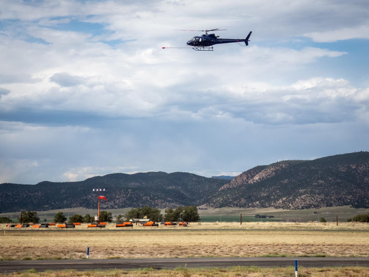 The image features a helicopter flying over a open area at an airport with mountains in the background. The helicopter is positioned towards the top center of the scene