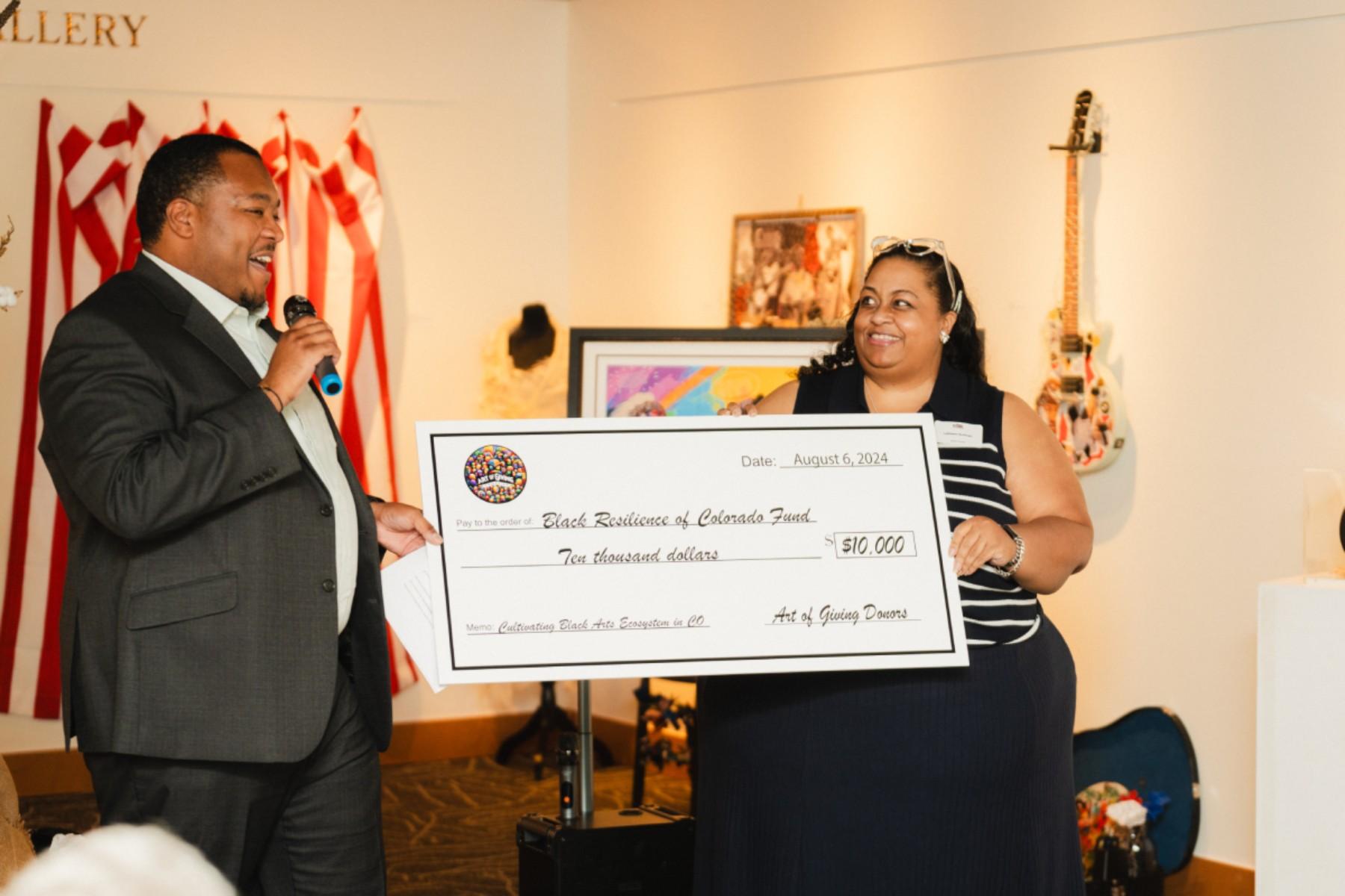 Image shows a man in a suit with a microphone on the left next to an oversized check being held by a woman standing behind the check to the right.