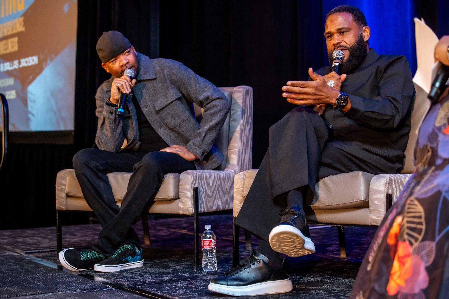 Photos shows two men on a stage sitting in casual chairs. They are both holding microphones and the man on the right has his legs crossed. He is speaking to an audience and gesturing with his arm.