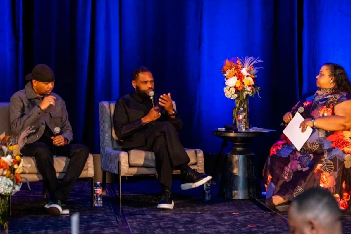 Photo shows three people seated in chairs on a stage with a bright blue backdrop. There is a vase of flowers next to the two men on the left between them and the woman on the right.