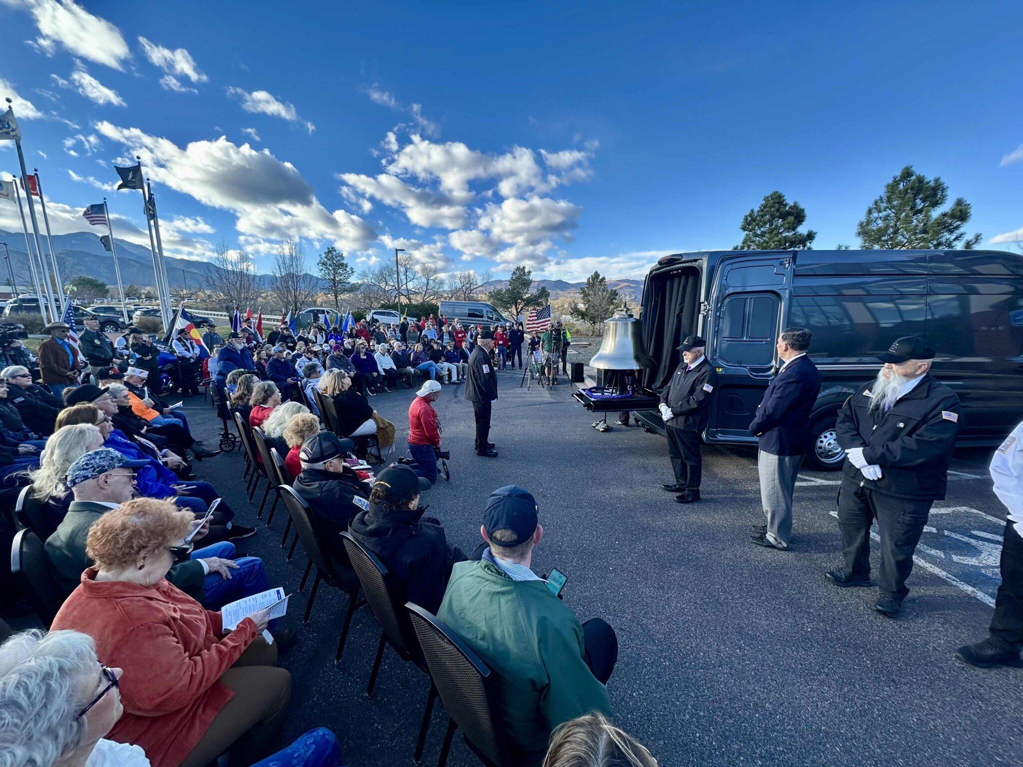 A crowd is seated for an outdoors dedication ceremony of a large bronze bell.