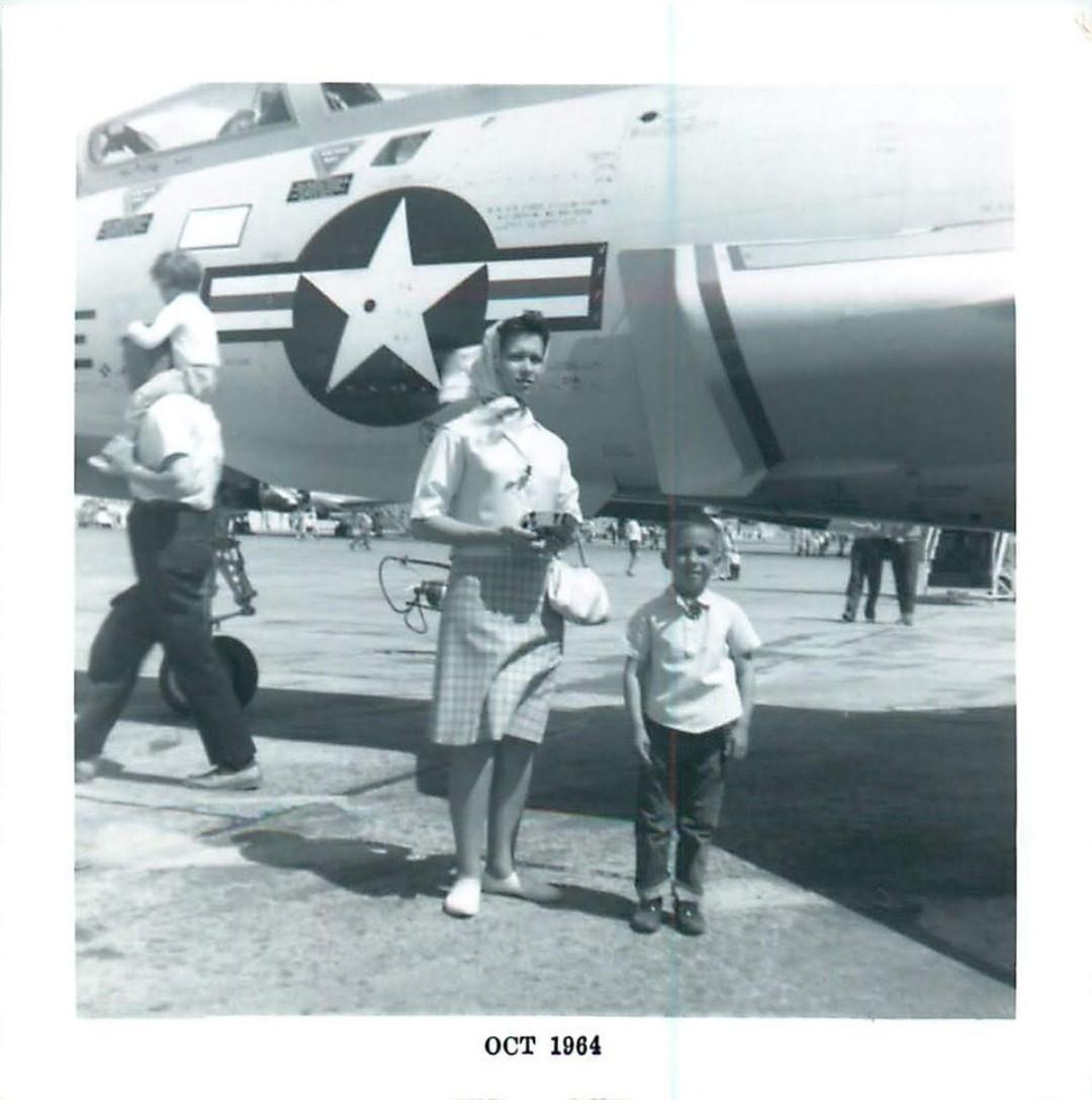Black and white photo shows a woman in a checkered dress and headscarf standing next to a small boy in front of a U.S. Air Force fighter jet.