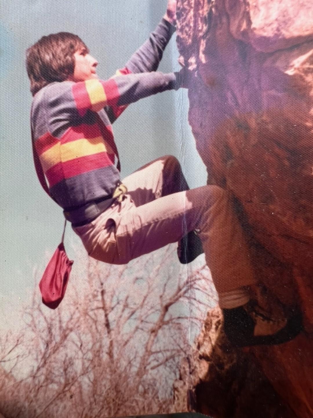 Image is a vintage photo circa 1978 showing a young man with a red, yellow, and blue striped shirt climbing a rockface in Garden of the Gods. His arms are outstretched and his legs are bent in a seated position.