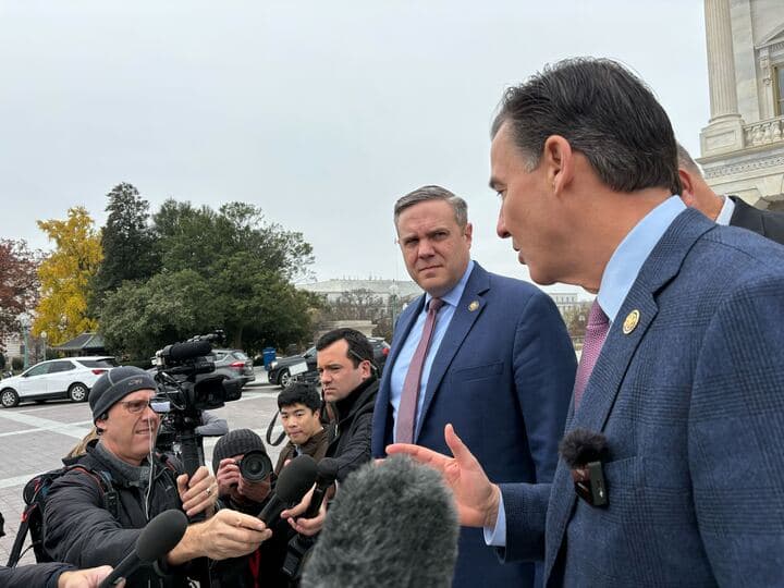A man in a suit watches another man in a suit speak to a crowd of people behind TV cameras