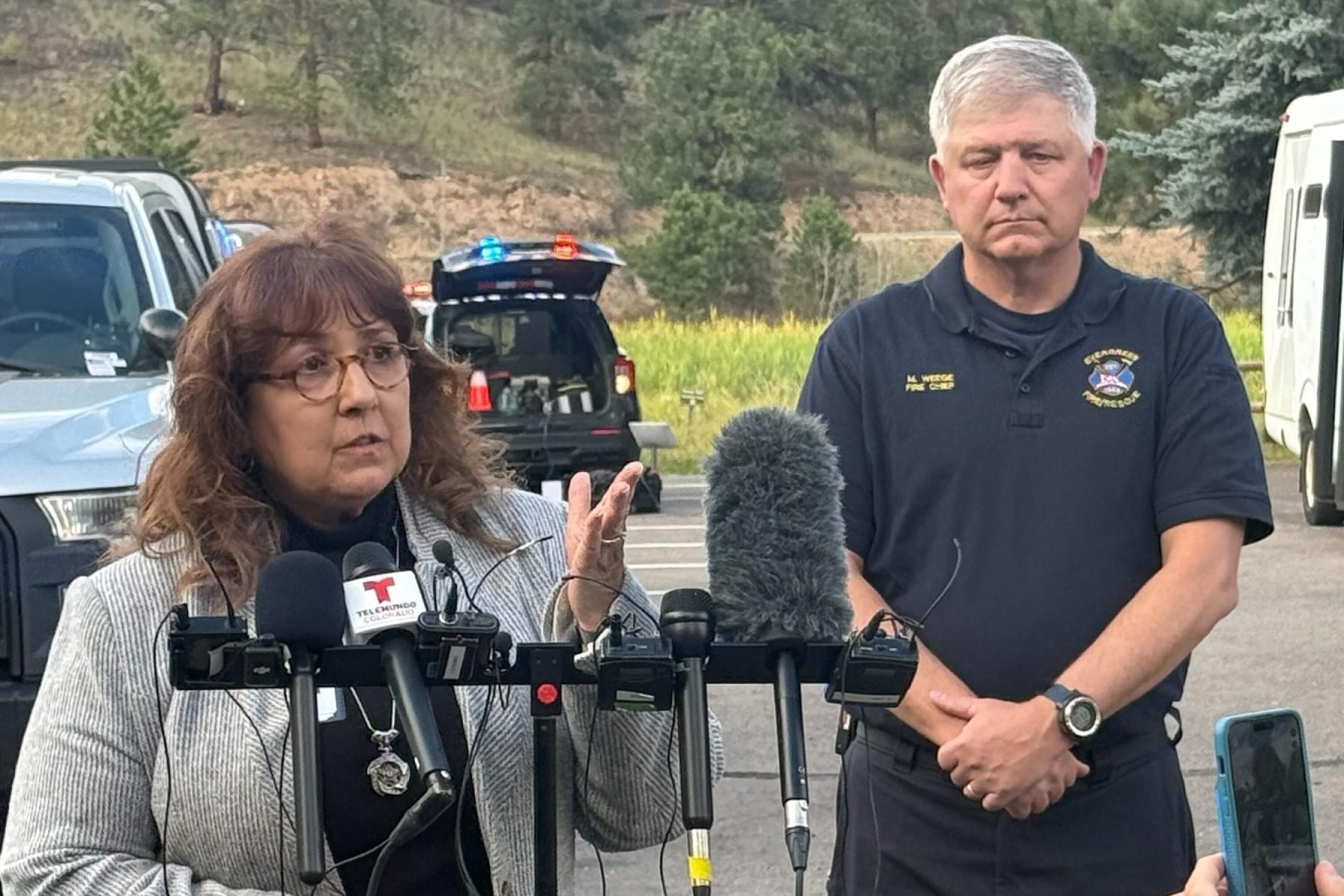A woman speaks in front of a microphone array. She has glasses on and wears a gray cardigan over a black shirt and pants. Standing behind her is a man with grey hair in a firefighting polo. The two are addressing the press in a parking lot with trees in the background.