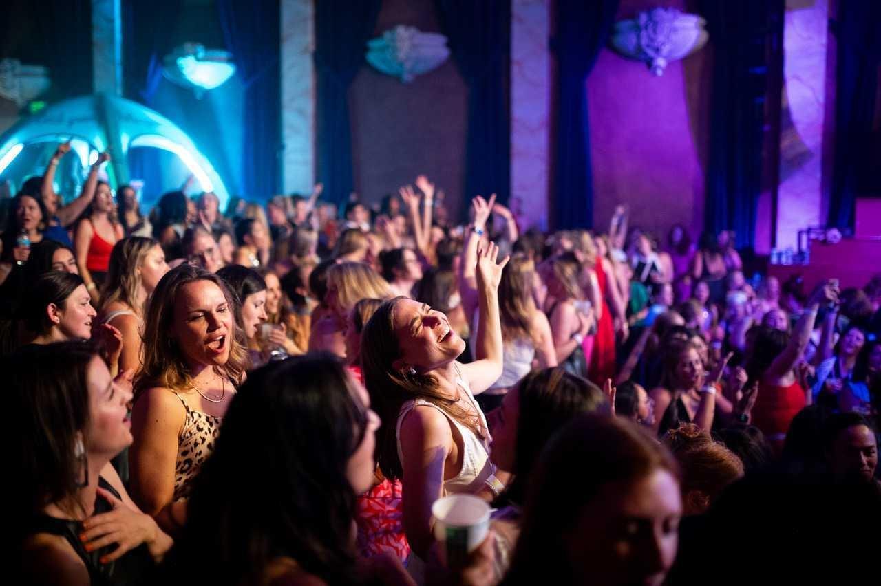 Photo of a crowd of women packed into a theater some with their arms up dancing.