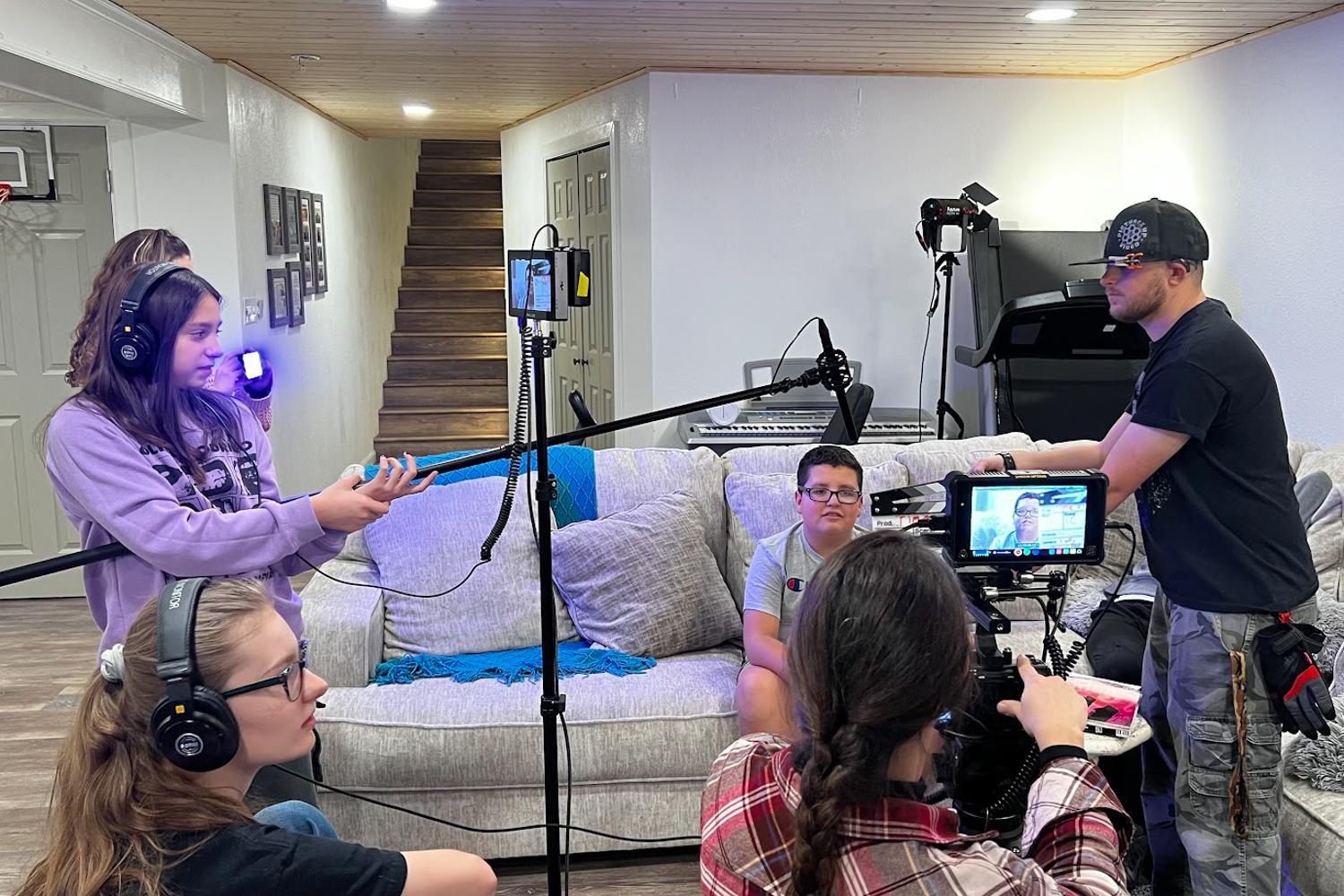 A group of young people are gathered in a living space using audio and visual recording equipment, with some sitting on a couch and others standing.