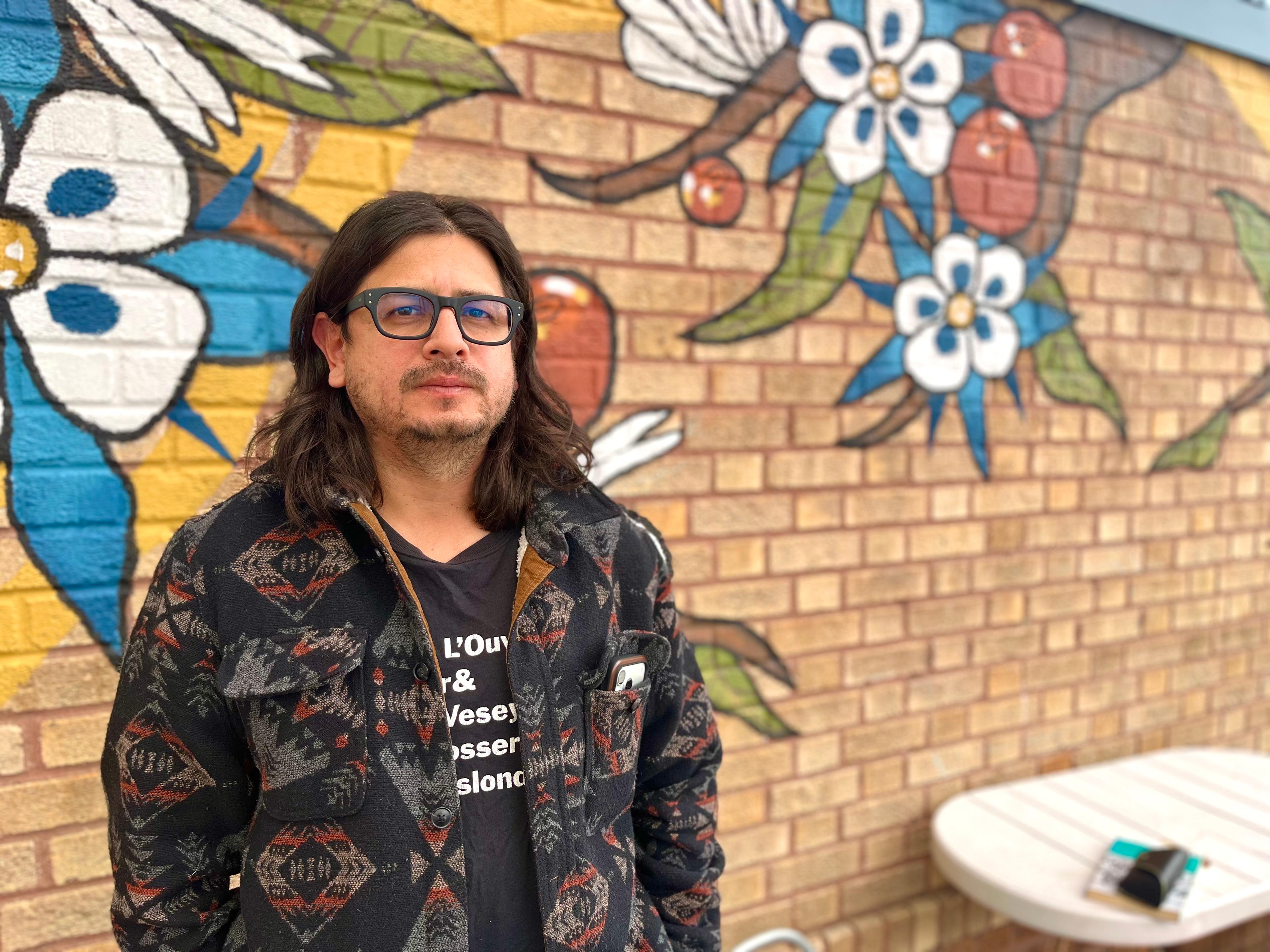 Director Raúl O. Paz Pastrana stands in front of a brick wall painted with columbine, leaves, and red berries.