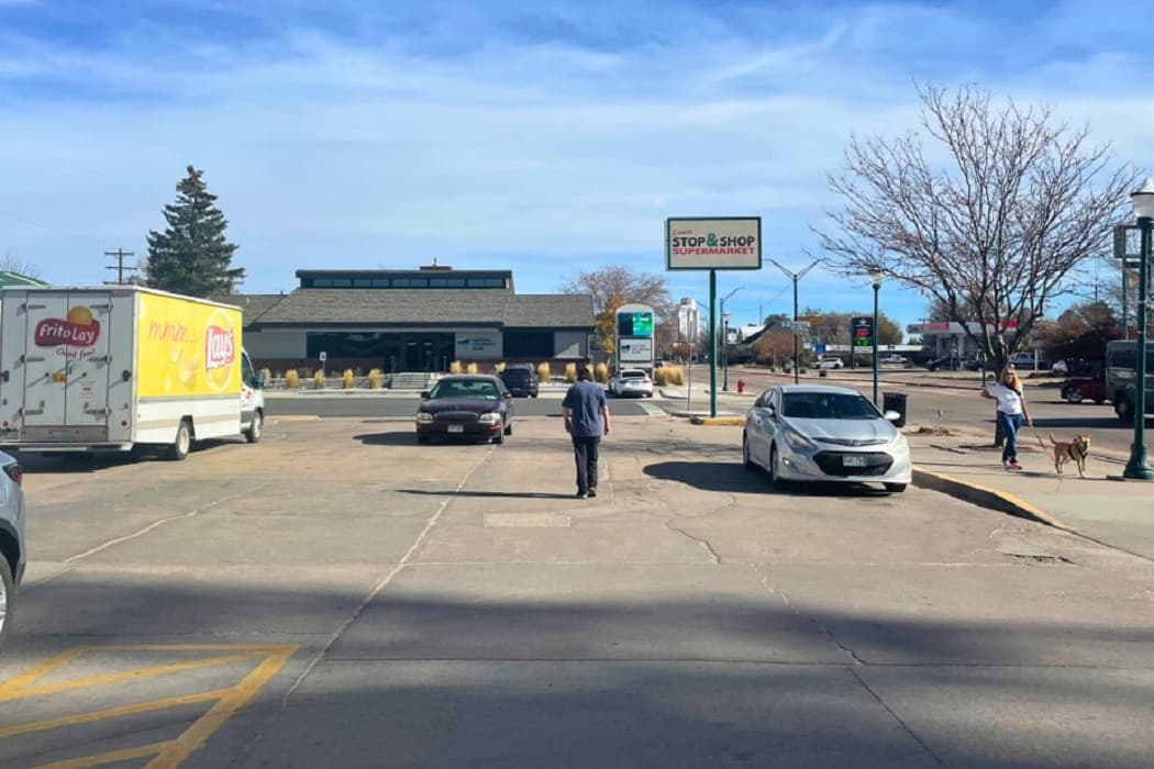 A person is walking in the parking lot of a store. There are cars parked along the street, and a truck is also present.