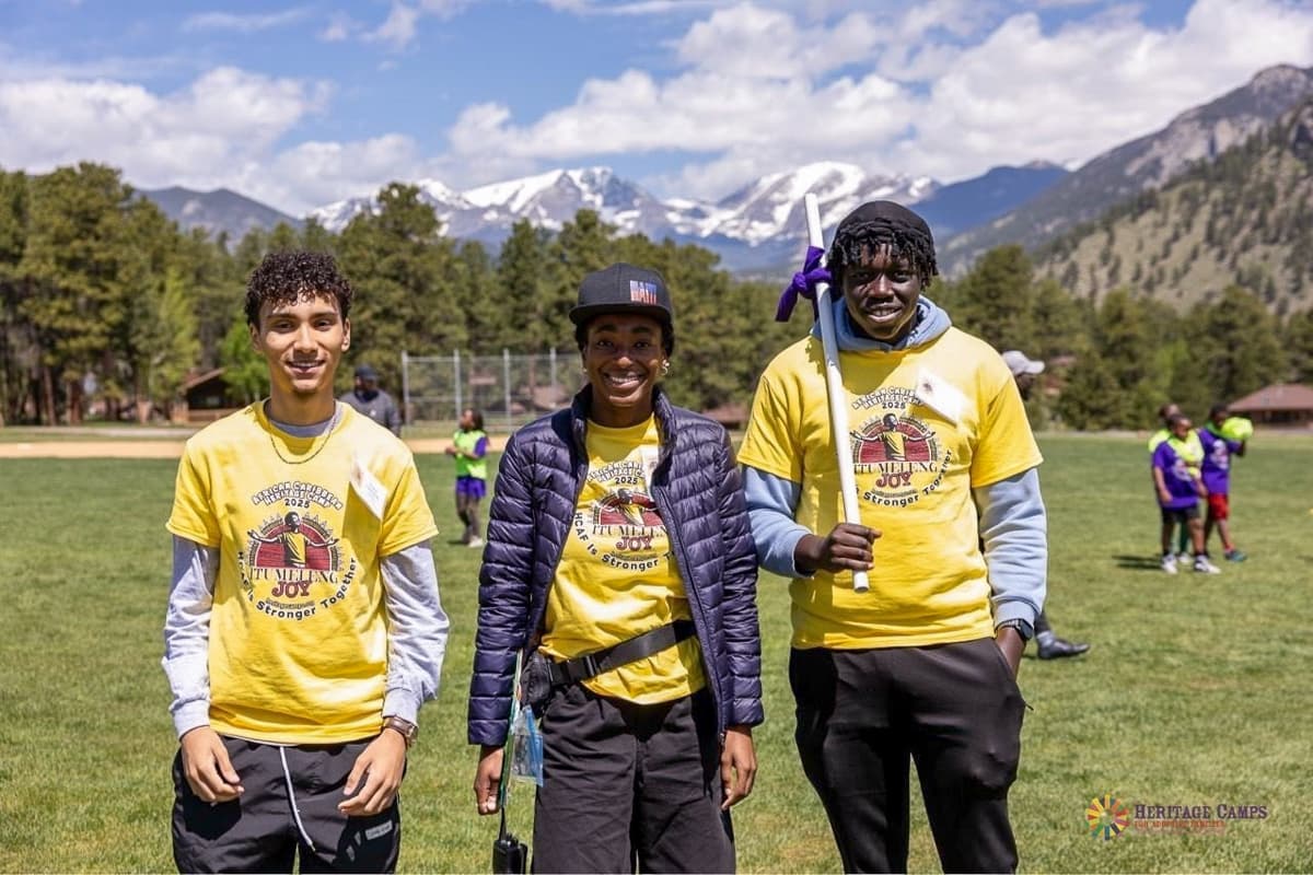 Photo shows three teenagers in yellow tee shirts standing in front of a baseball field. The boy on the right is holding a white stick with a purple ribbon tied on the end.
