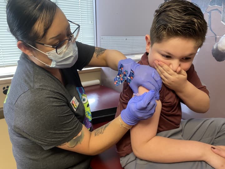 A woman is giving a young boy a vaccine. She is wearing a mask and surgical gloves. The boy is holding one hand over his mouth as he receives the shot.