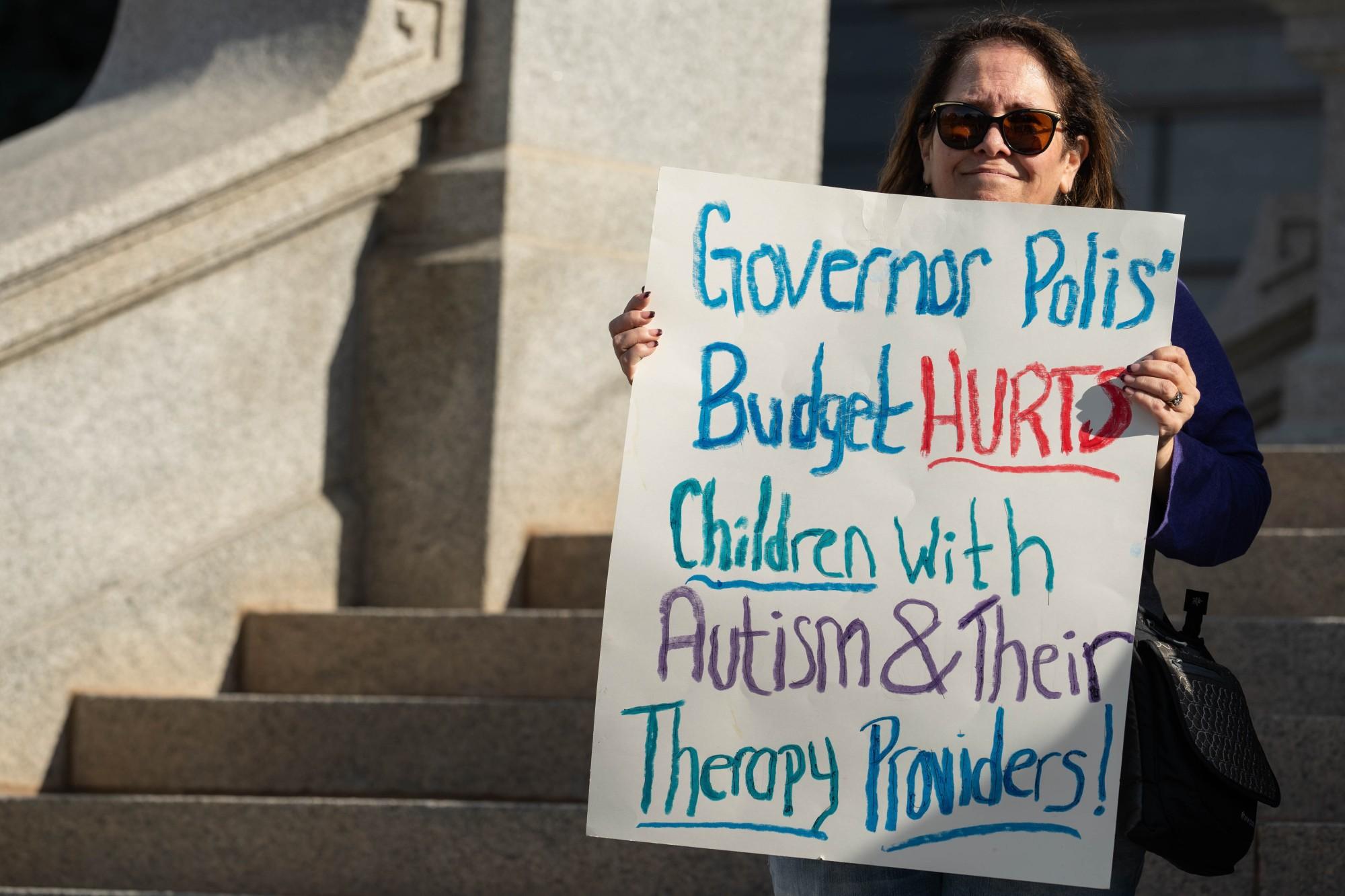 A woman in sunglasses stands on the Capitol steps with a handwritten sign reading &quot;Governor Polis' budget hurts children with autism and their therapy providers!&quot;