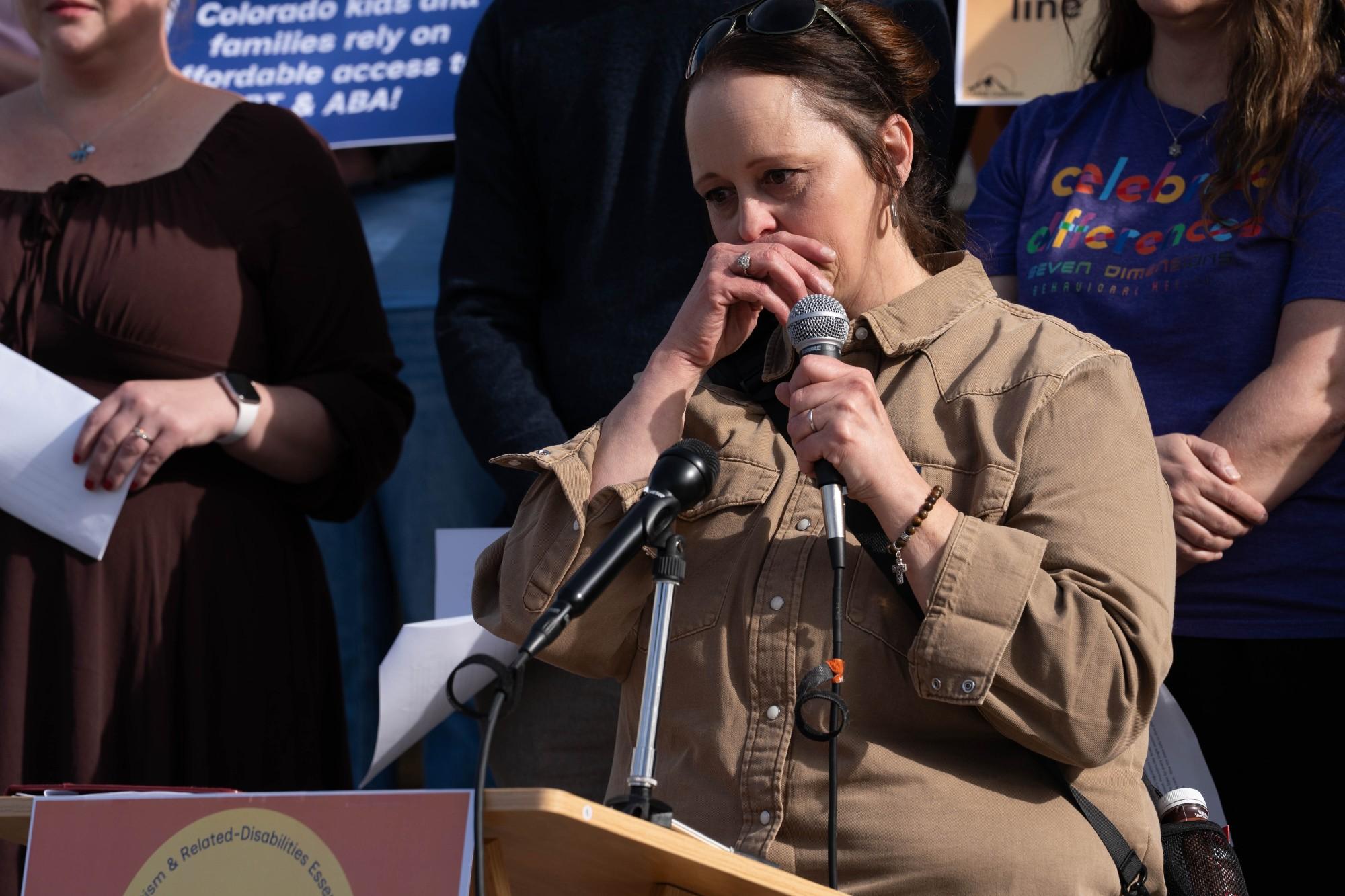 A woman in a brown shirt looks sad and covers her mouth as she speaks into a microphone at a podium with a crowd behind her.