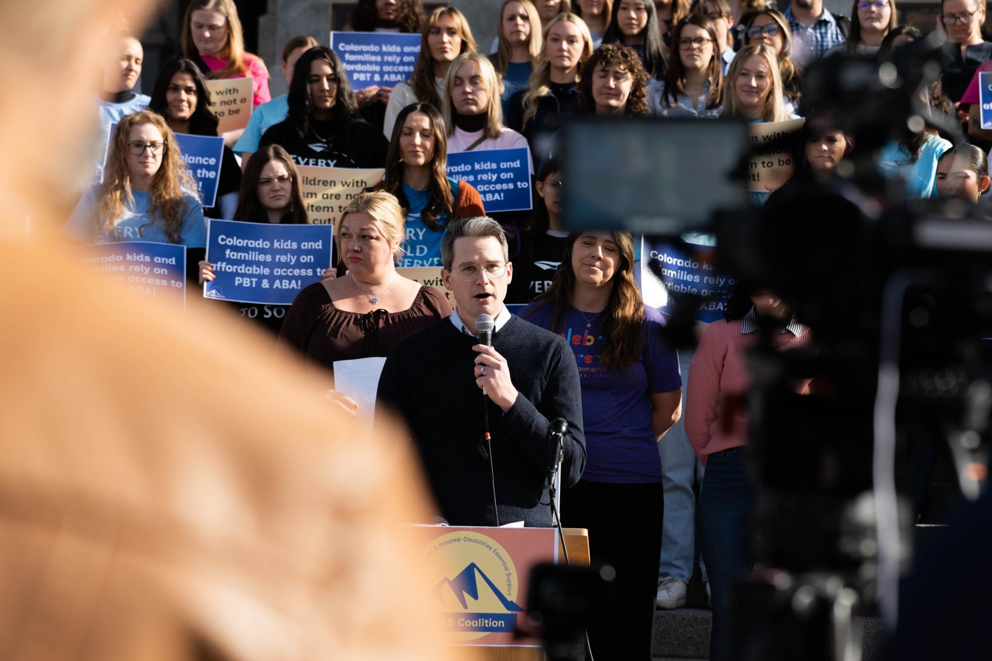 A man in a blue sweater speaks into a microphone with a crowd holding signs behind him on the state capitol steps