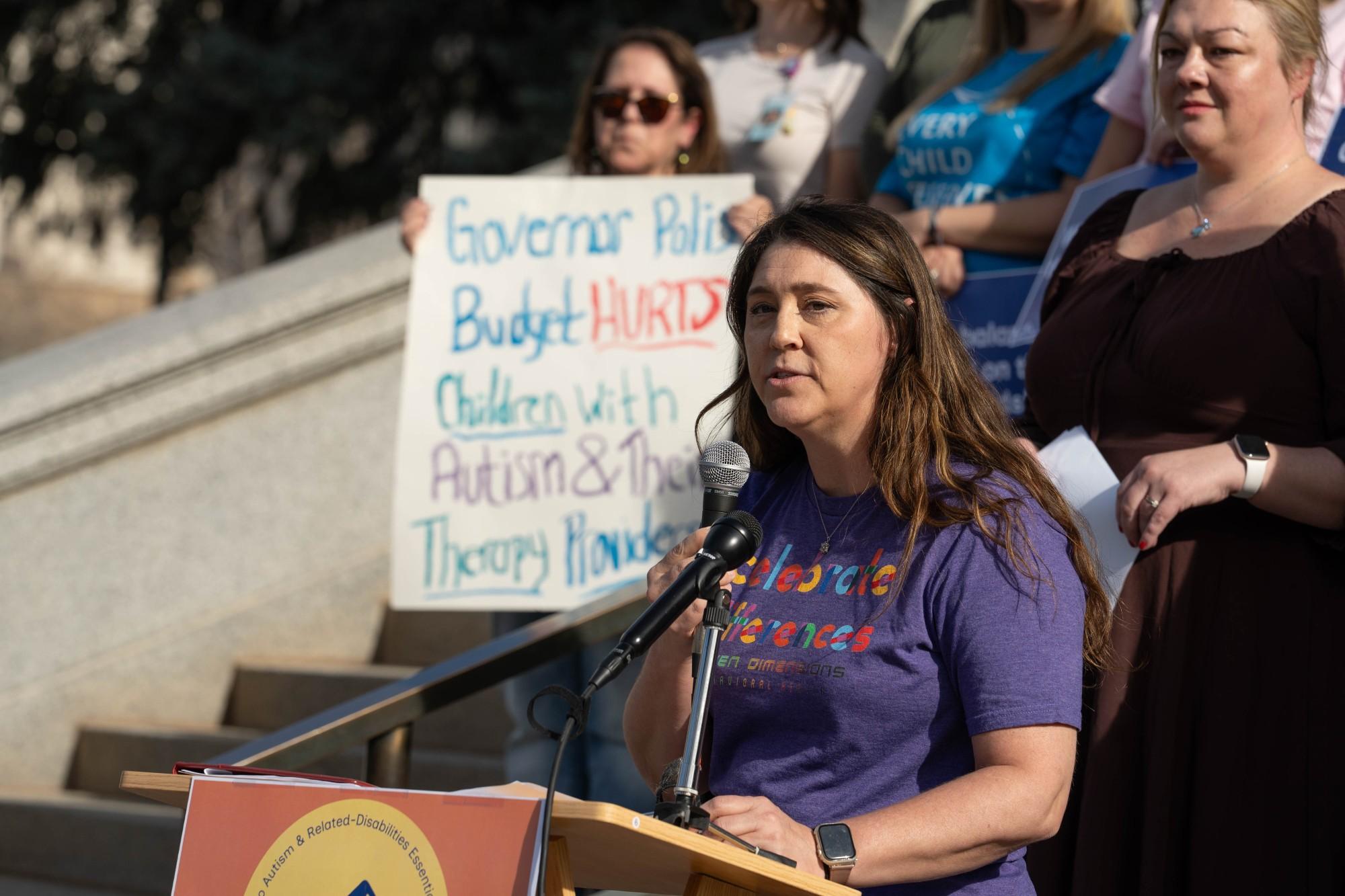 A woman in a blue shirt speaks into a microphone at a podium with a crowd behind her