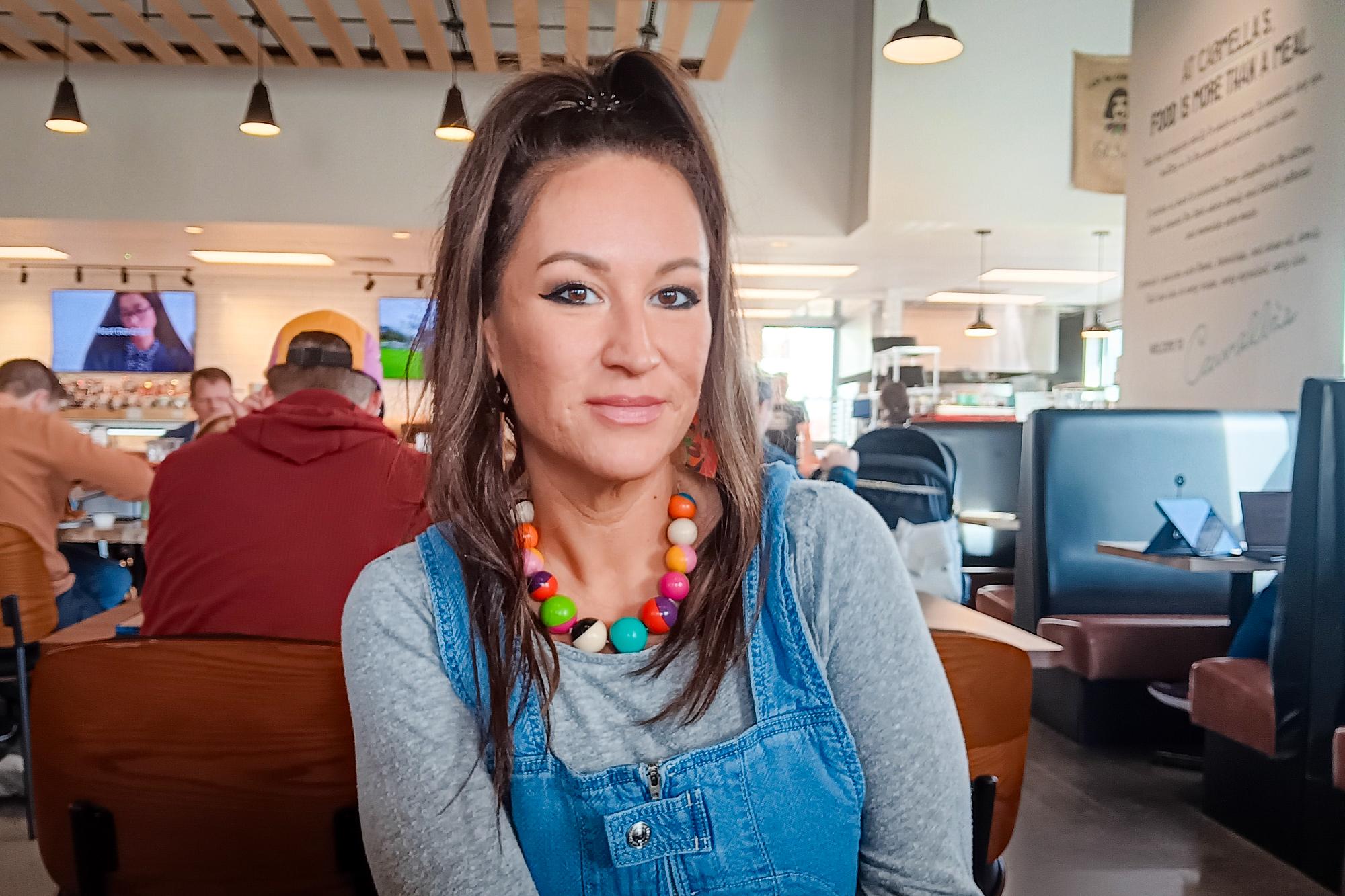 A person in a white top grey undershirt looks at the camera sitting in a booth