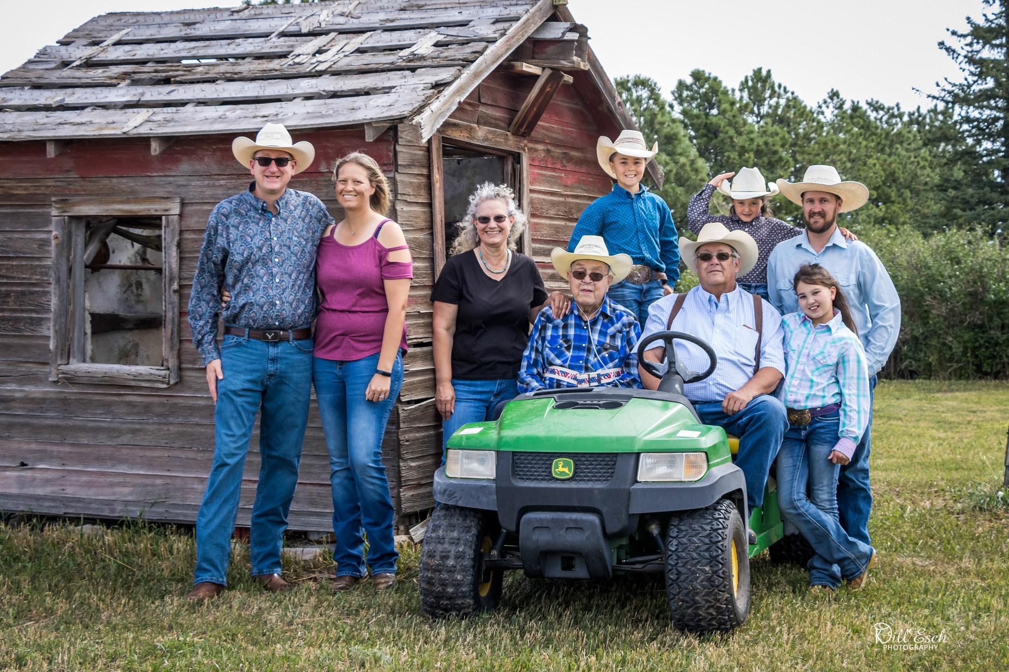 ›A multigenerational family wearing cowboy hats poses outdoors beside a weathered wooden shed. Two older adults sit on a green utility vehicle at center, surrounded by adults and children standing close together on grass, with trees in the background.