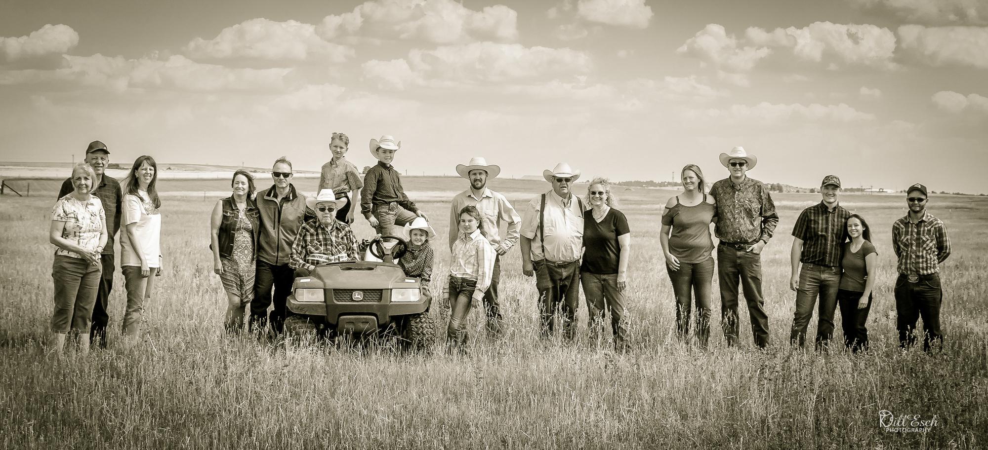 A wide, black-and-white portrait shows a large, multigenerational family standing in a grassy field under a partly cloudy sky. Adults and children line up shoulder to shoulder, some wearing cowboy hats, with two older family members seated on a utility vehicle near the center of the group.