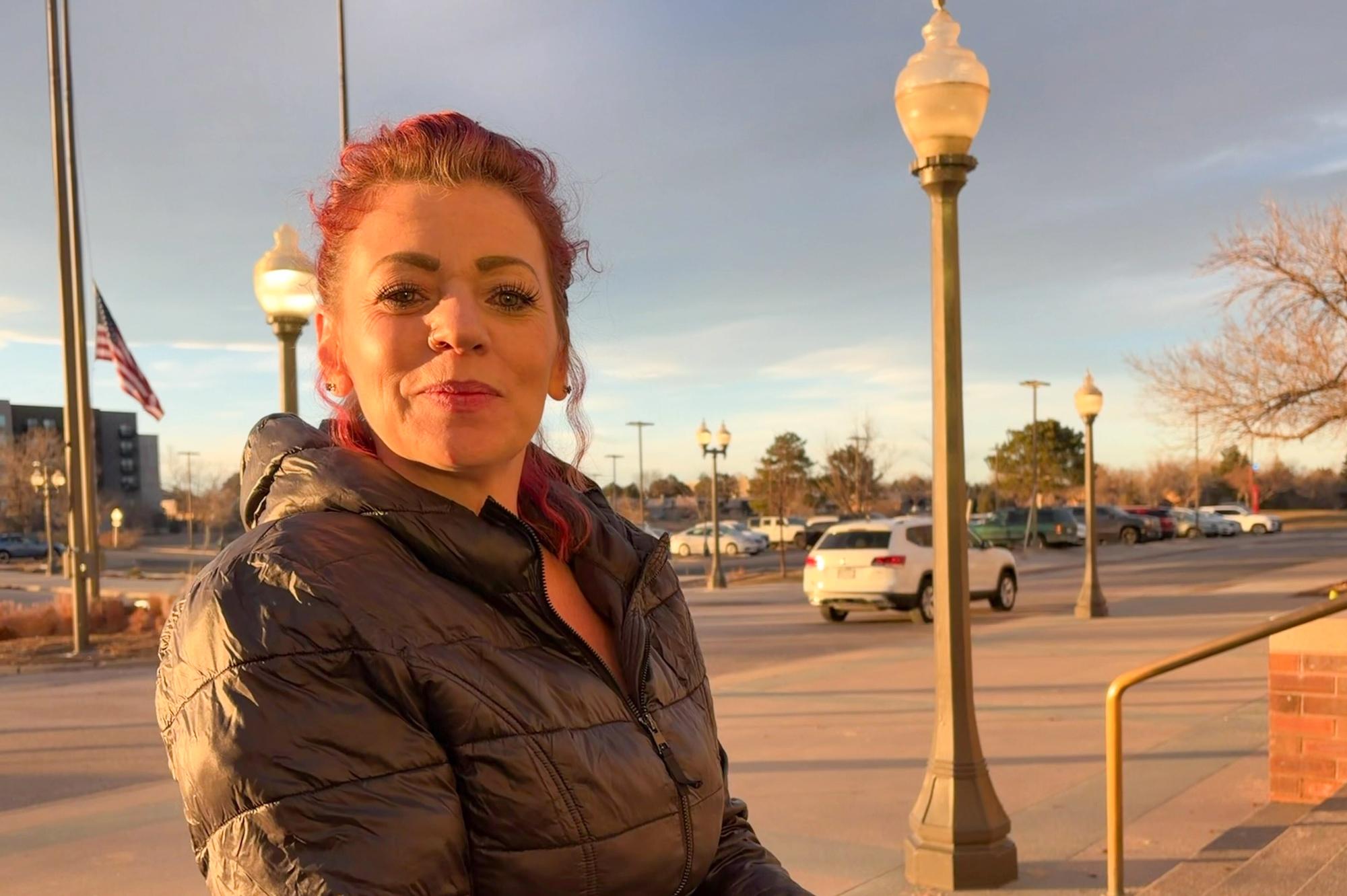 A woman with red hair is standing in a parking lot, smiling at the camera. She is wearing a black jacket and looking directly at the camera.
