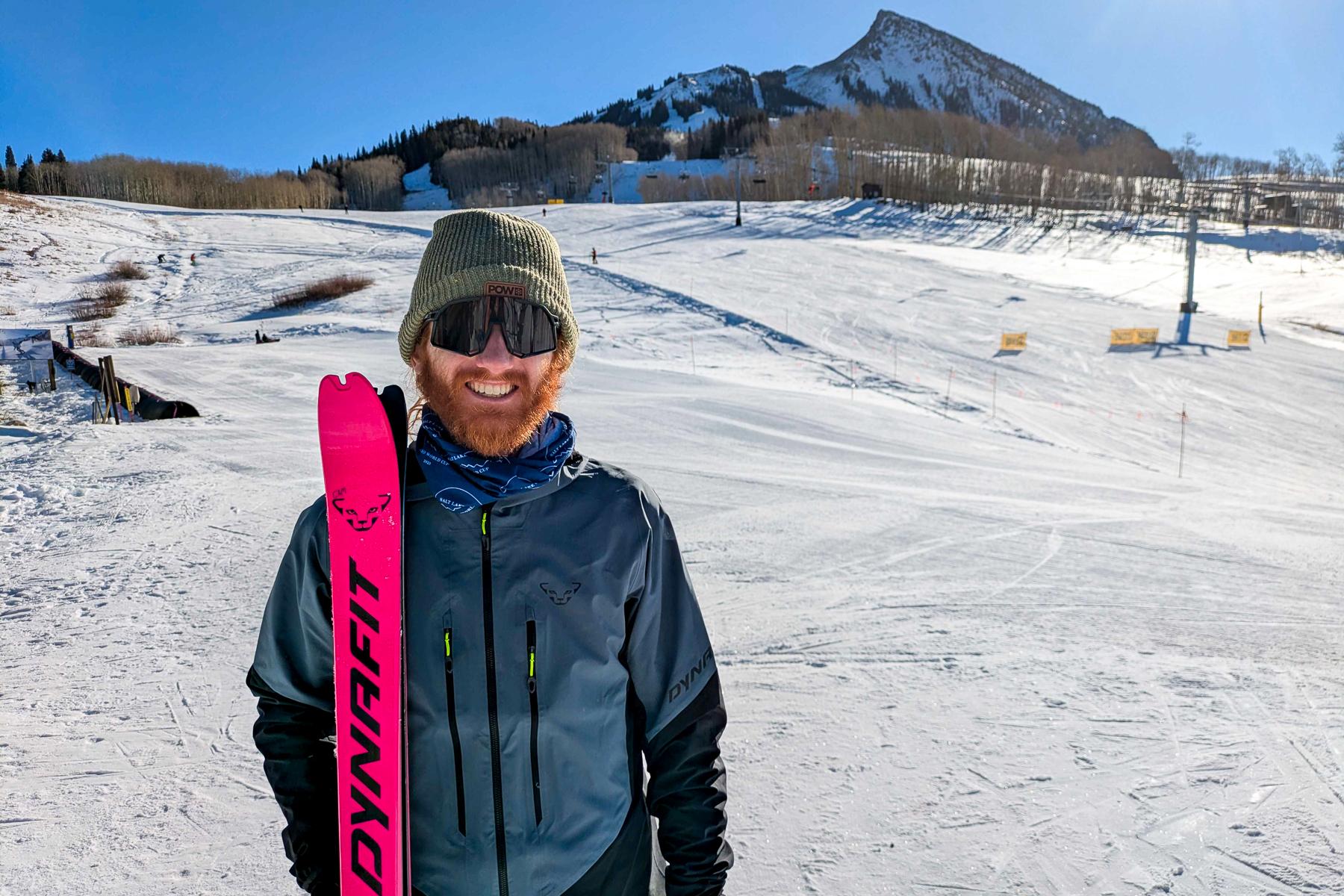 Cam Smith stands on a ski slope holding a pair of florescent skis and wearing a green stocking cap with a teal ski jacket.