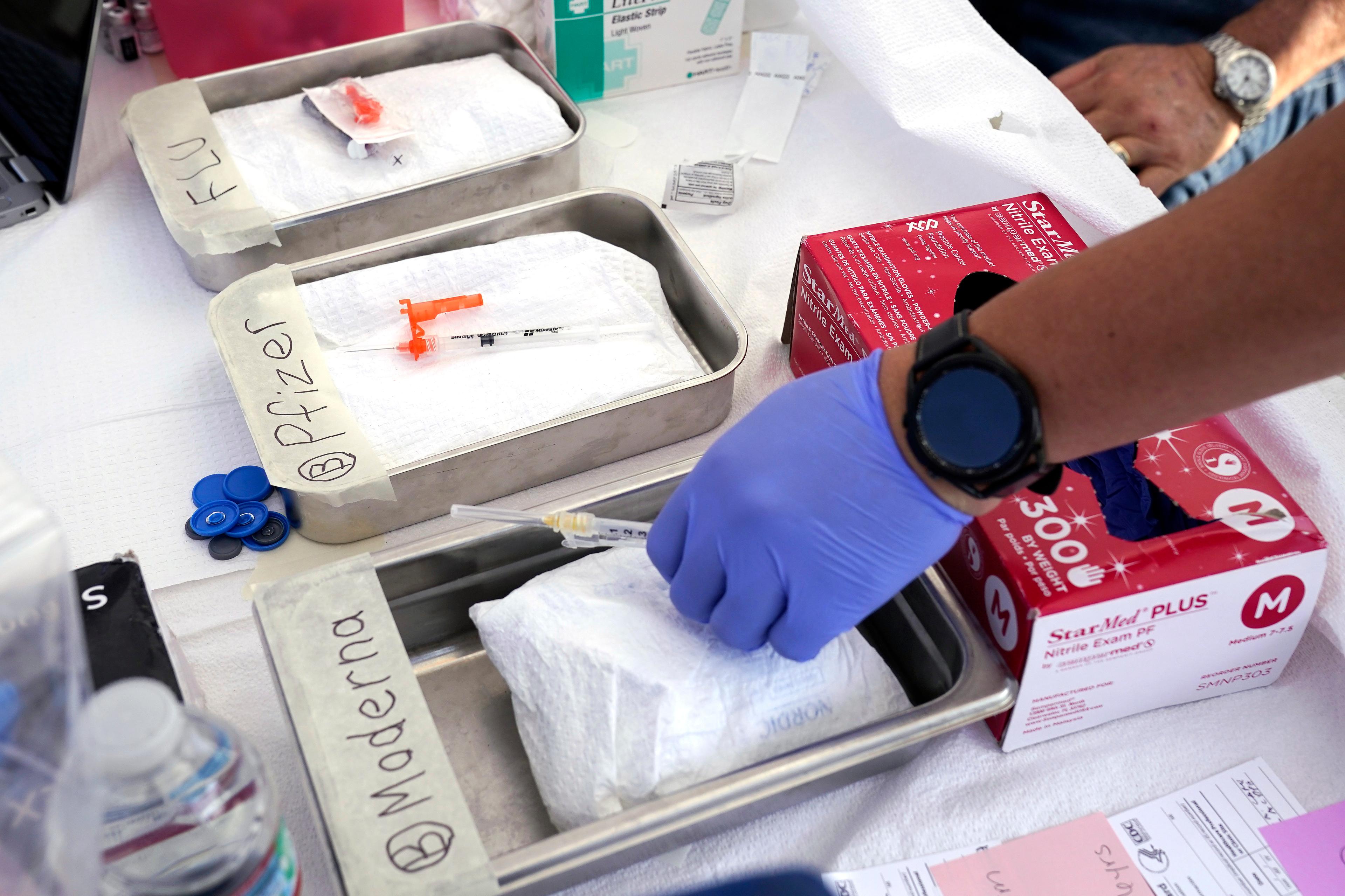 Syringes with vaccines lie in trays marked "Moderna," "Pfizer" and "Flu." A blue-gloved hand reaches for a Moderna syringe.