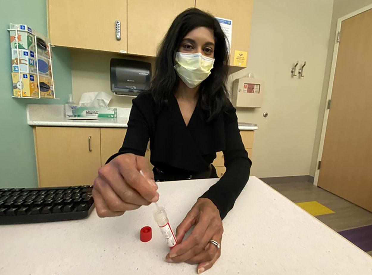 A woman wearing a face mask is holding a vial of liquid, possibly a medical sample. She is standing in a room with a counter and a keyboard nearby.