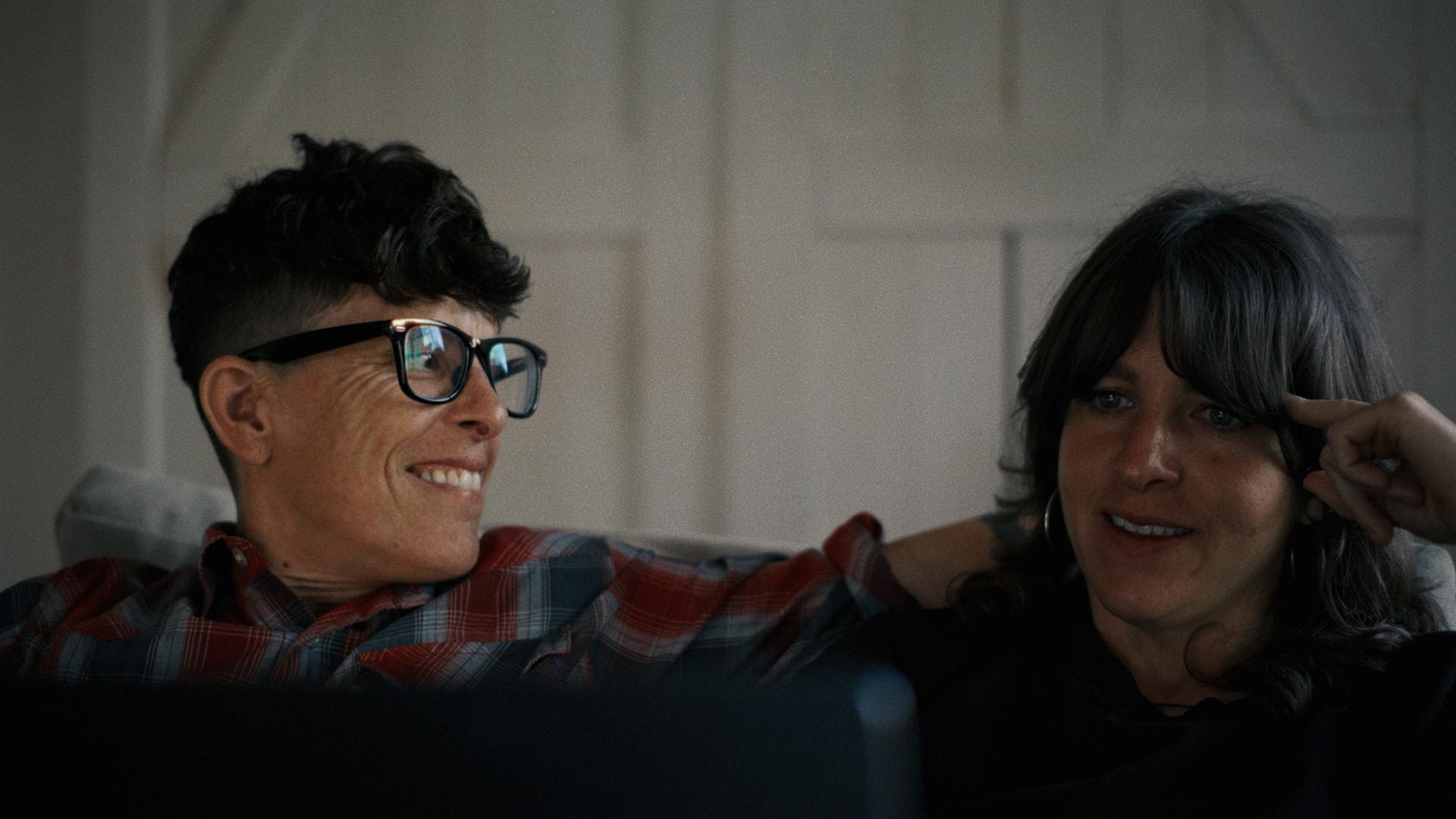 Image shows the heads of two people. On the left is a person with short black hair and black glasses, smiling and looking toward the person on the right, who has long black hair.