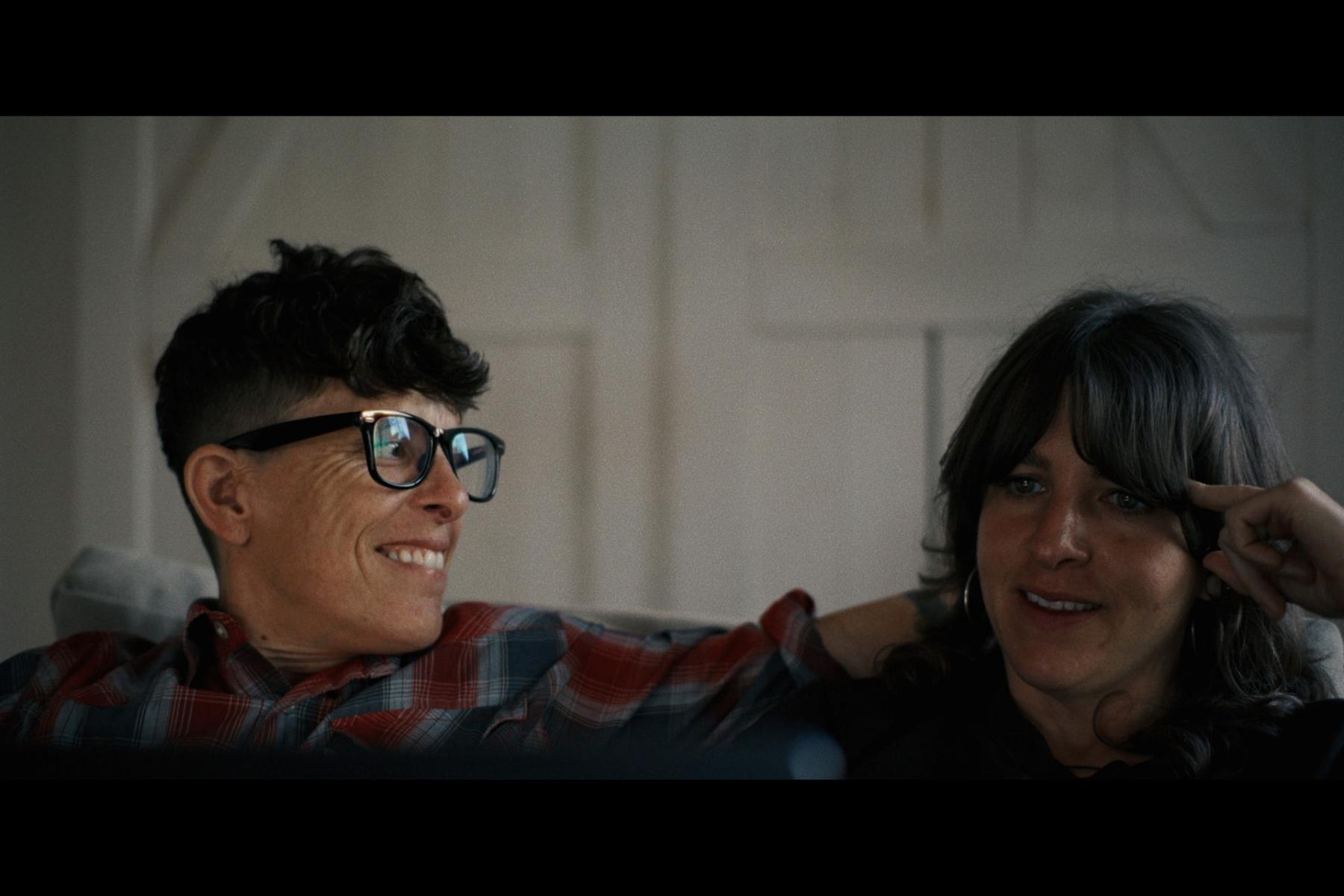 Image shows the heads of two people. On the left is a person with short black hair and black glasses, smiling and looking toward the person on the right, who has long black hair.