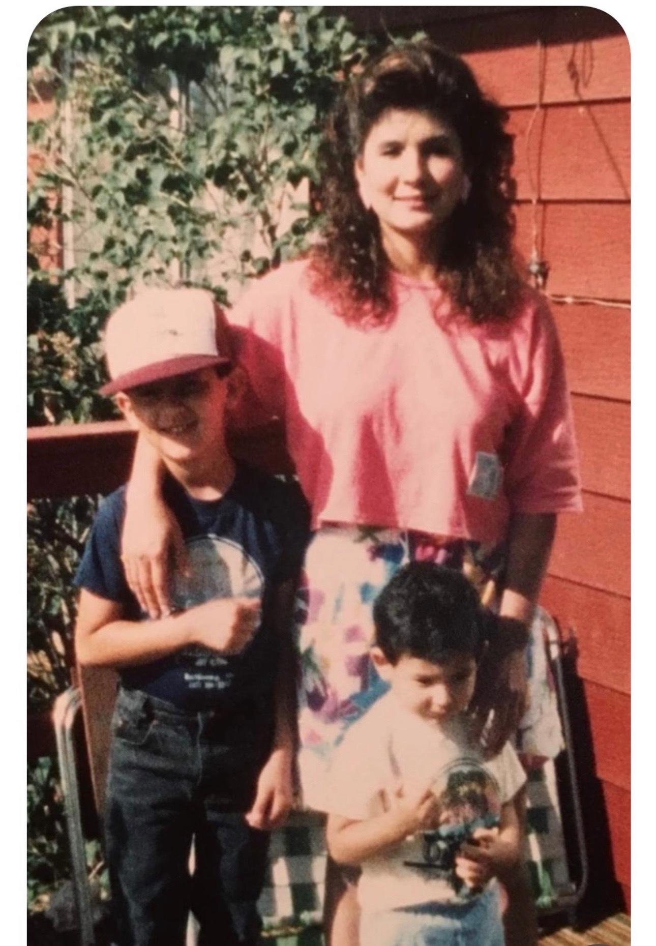 A vintage, film photo of Democratic House Majority Leader Monica Duran with her two young sons standing in front of her. Her arm is aroud the older boy, who is wearing a baseball cap. The younger boy is holding a toy.