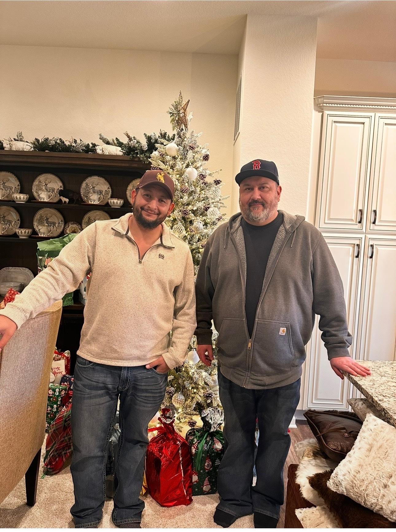 Patrick Ellis (left) and David Duran (right) stand in a living room in front of a Christmas tree.