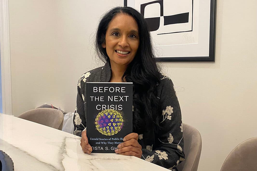A woman is sitting at a table, holding a book titled "Before the Next Crisis." She is smiling at the camera.