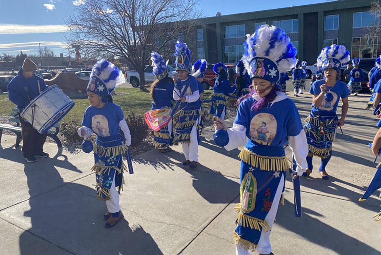 A group of people wearing colorful costumes and headdresses are walking down a sidewalk. The participants are spread out along the sidewalk, with some closer to the foreground and others further back.