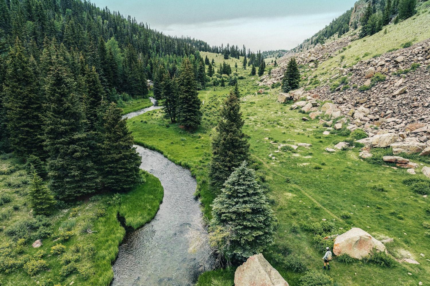 A small river flows through a forested valley, large rocks are on one side and evergreen trees on the other