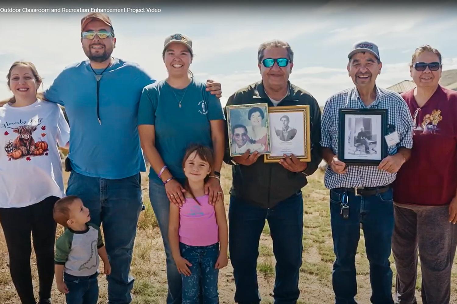 A group of people, including two young children are posing for a picture. Two hold photos from previous eras