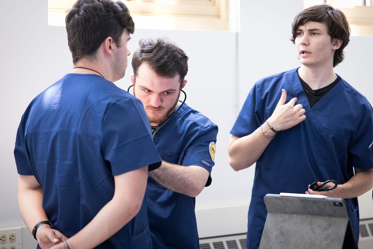 Three men in blue shirts are standing in a room, engaged in conversation. One of the men is wearing a stethoscope, indicating that they might be discussing medical topics or sharing their experiences in the field.