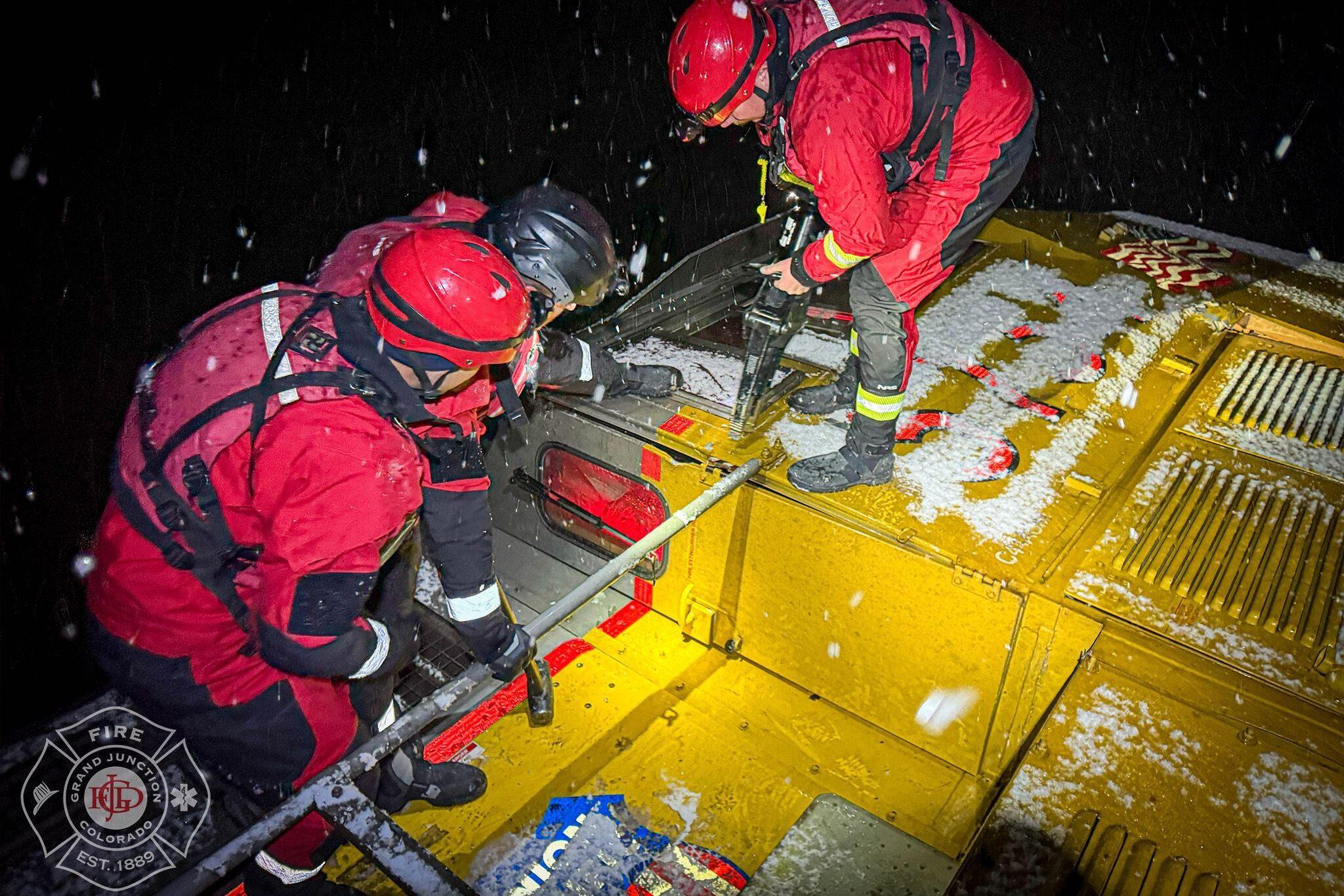 Three firefighters are working together to access a derailed train. The firefighters are wearing red jackets and helmets, and they are equipped with tools.