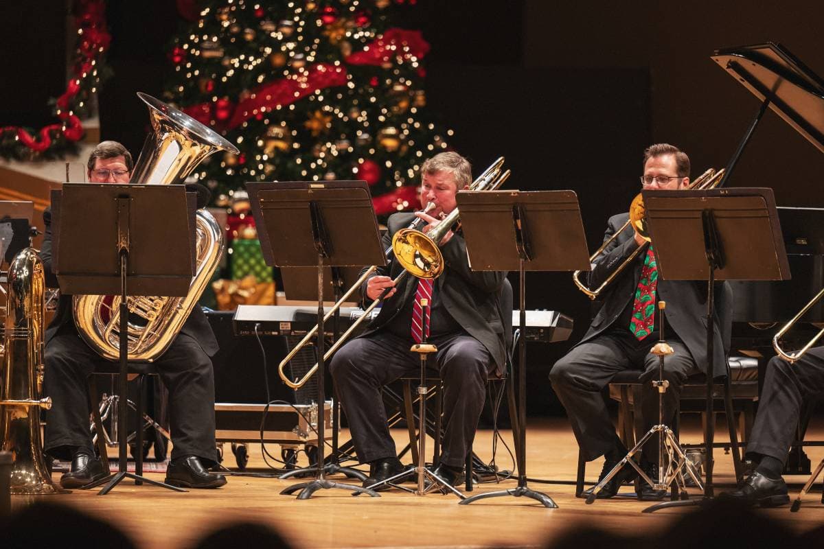 Two people playing trombones, one playing a tuba in front of a Christmas tree.