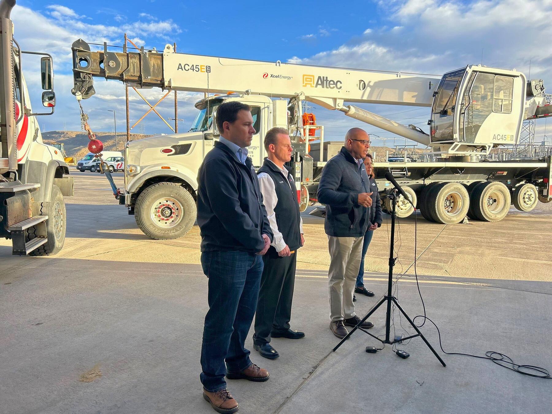 A group of people standing in front of a large crane truck with one person speaking into a standup microphone.