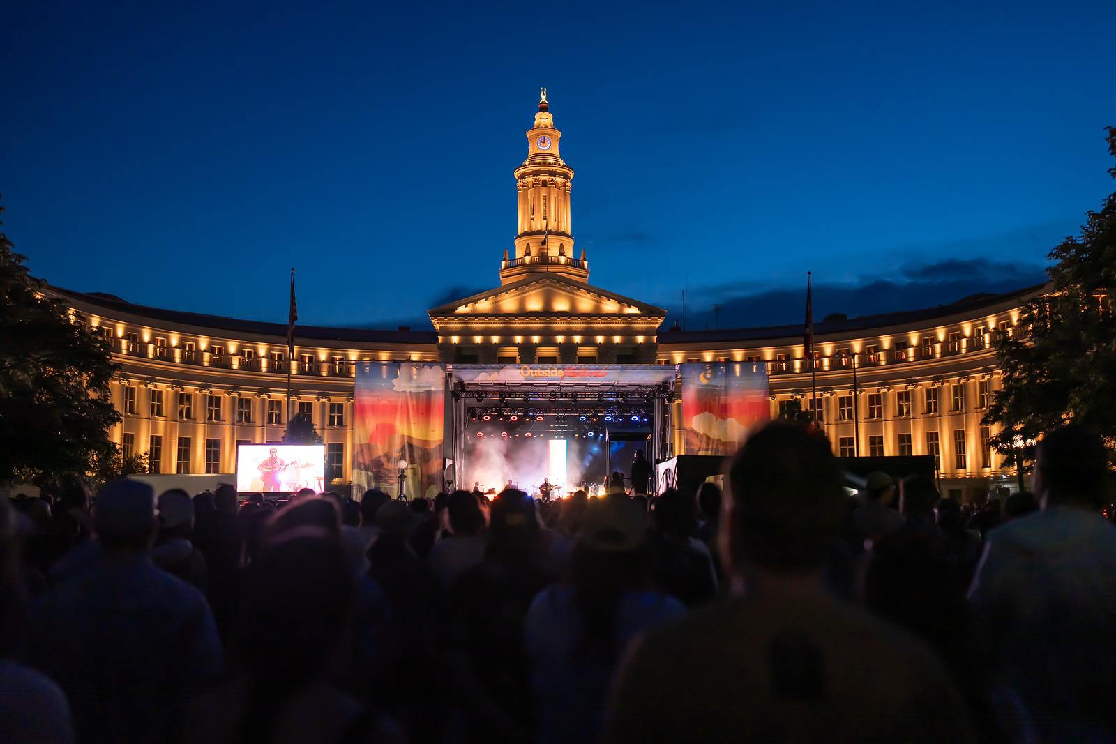 A large crowd watches a music festival stage, with an illuminated large government building behind the stage.