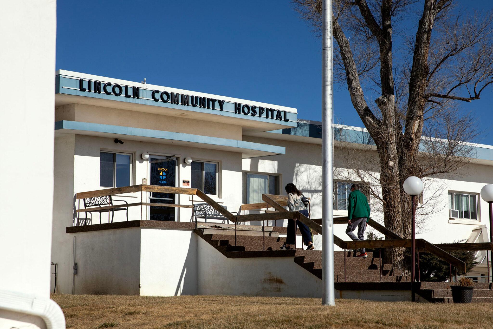 A squat white building is adorned with lettering reading "LINCOLN COMMUNITY HOSPITAL." Two people walk up stairs towards a door.