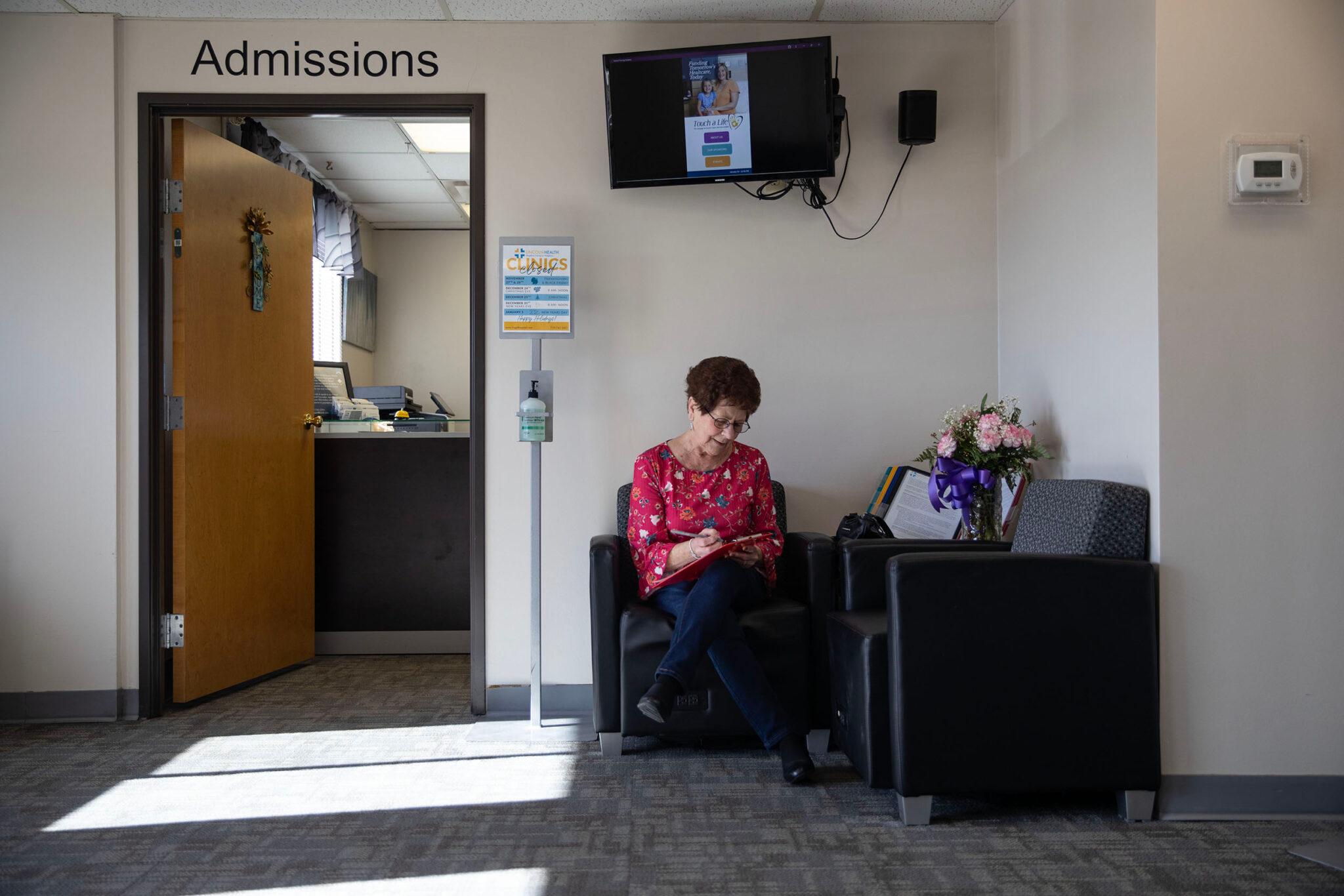 A woman in a red blouse sits in a black leather chair next to a door labeled "admissions." She peers down to the clipboard she's filling out with a pen.